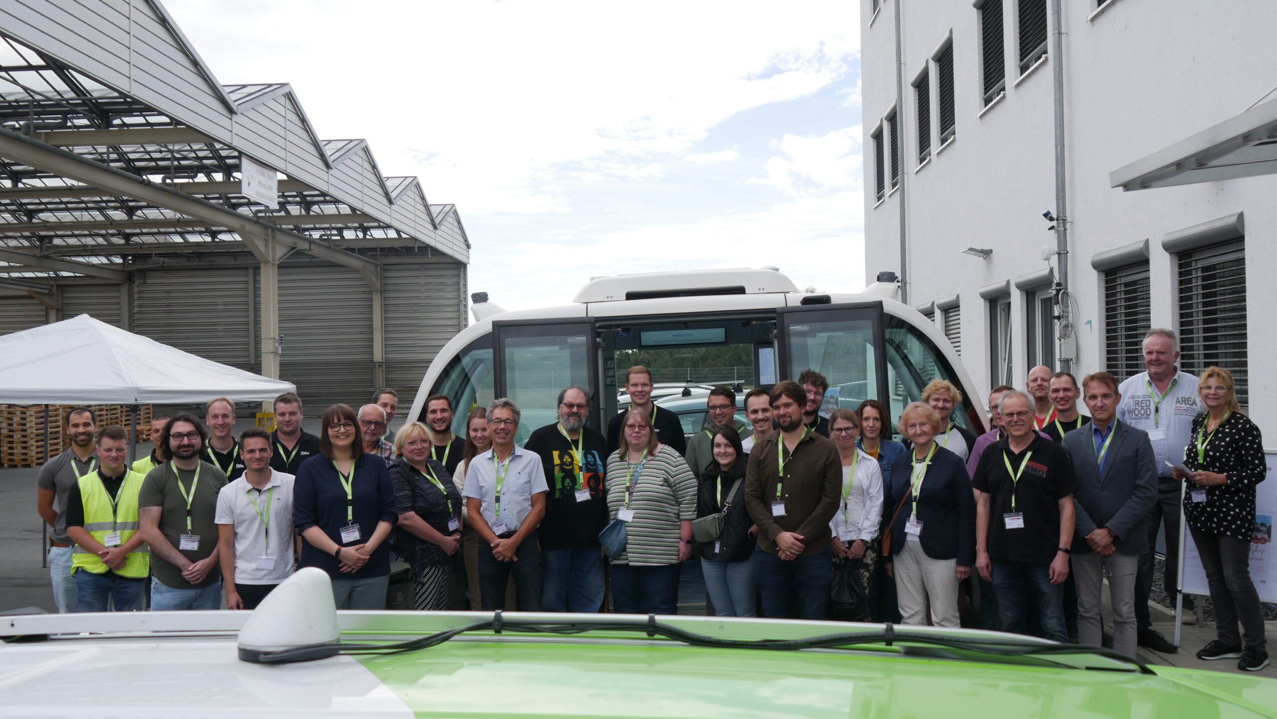 Eine Gruppe von Menschen steht vor einem weißen Shuttlebus und posiert für ein Foto an der Hochschule Coburg. Sie befinden sich in einem Industriegebiet, im Hintergrund sind ein weißes Gebäude und Metallkonstruktionen zu sehen. Die meisten tragen Lanyards.