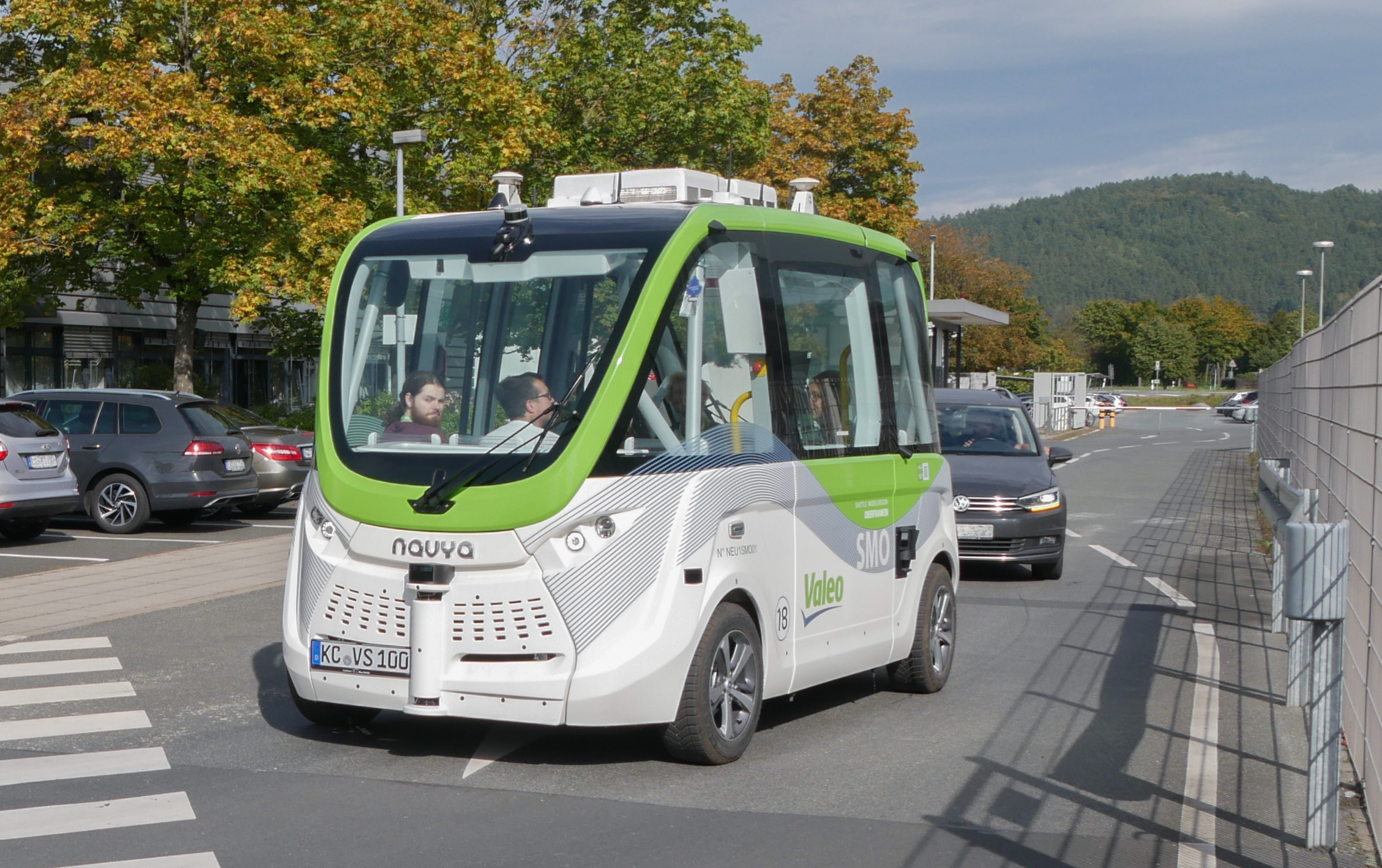 Ein kleiner fahrerloser Shuttlebus mit der Aufschrift „Navya“ fährt die Straße nahe der Hochschule Coburg entlang. Das weiße Fahrzeug mit grünen Akzenten transportiert mehrere Passagiere, während im Hintergrund Autos vorbeifahren und Bäume die Straße unter einem klaren Himmel säumen.