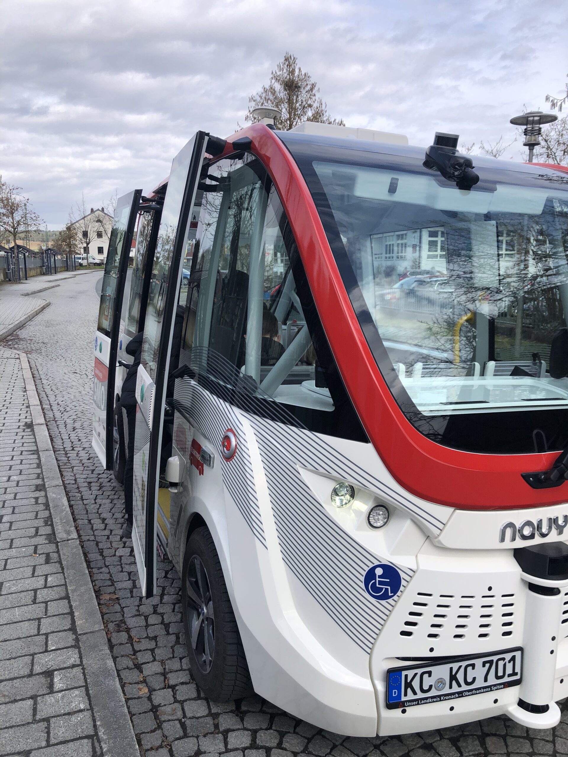 Ein kleiner, moderner autonomer Bus mit rot-weißer Außenfassade steht auf einer Kopfsteinpflasterstraße in der Nähe der Hochschule Coburg. Seine Türen sind geöffnet und an der Seite ist das Symbol für Rollstuhlzugänglichkeit zu sehen. Bäume und Gebäude säumen anmutig den Hintergrund.