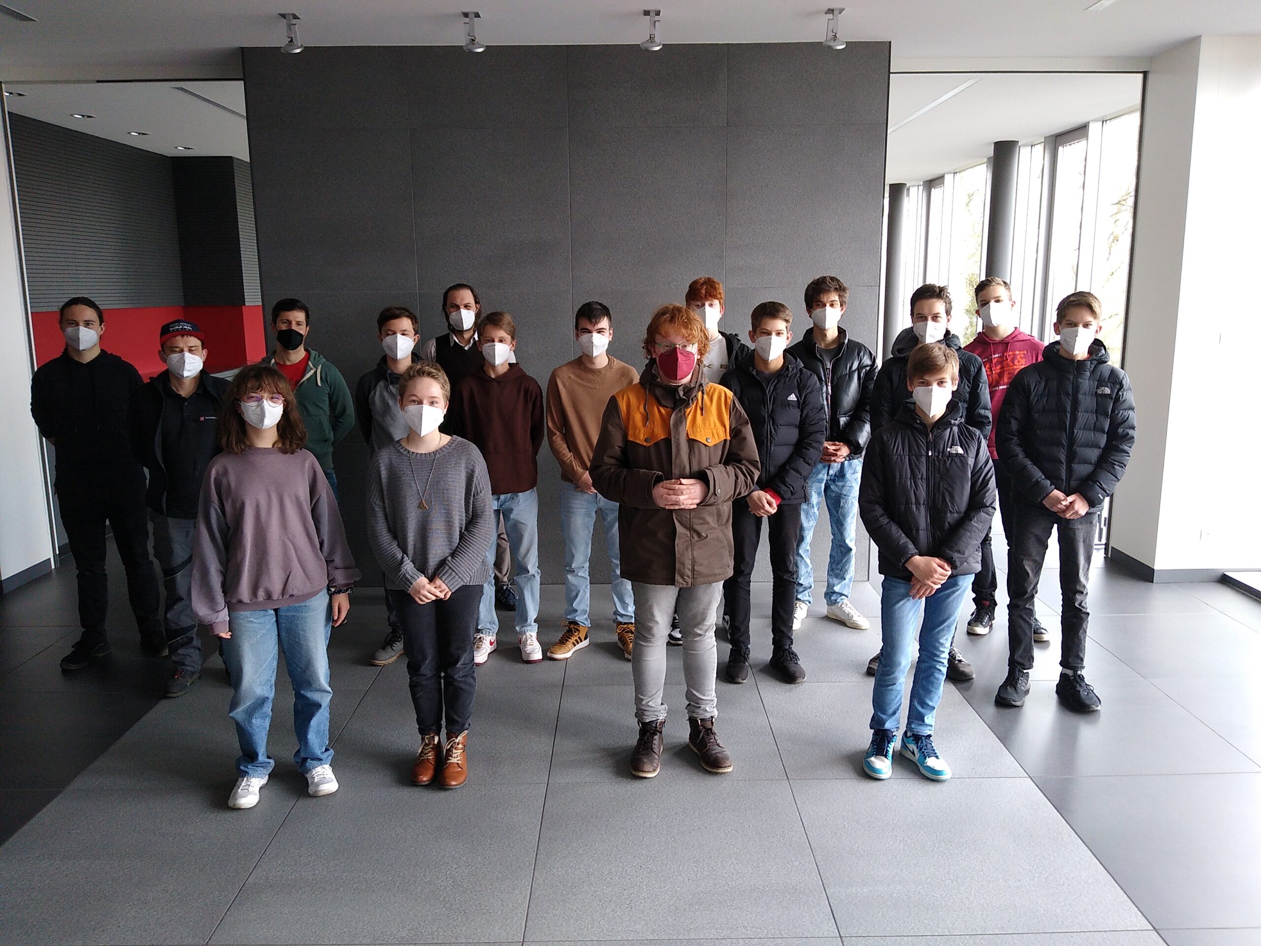 Eine Gruppe von Menschen mit Mund-Nasen-Schutz steht in einem Raum mit großen Fenstern und einer dunklen Wand der Hochschule Coburg. Sie sind leger gekleidet und scheinen für ein Gruppenfoto zu posieren.