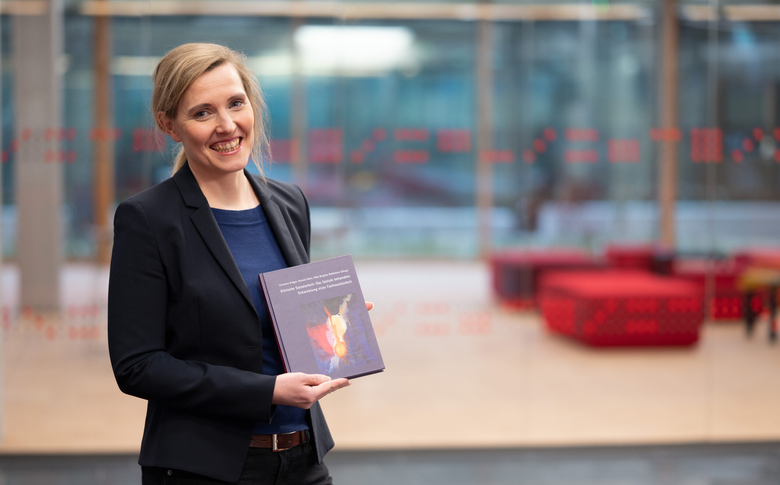 Eine Frau mit blonden Haaren lächelt und hält ein Buch in der Hand. Sie trägt einen schwarzen Blazer und steht in einem modernen, hell erleuchteten Raum mit den typischen roten Möbeln der Hochschule Coburg im Hintergrund.