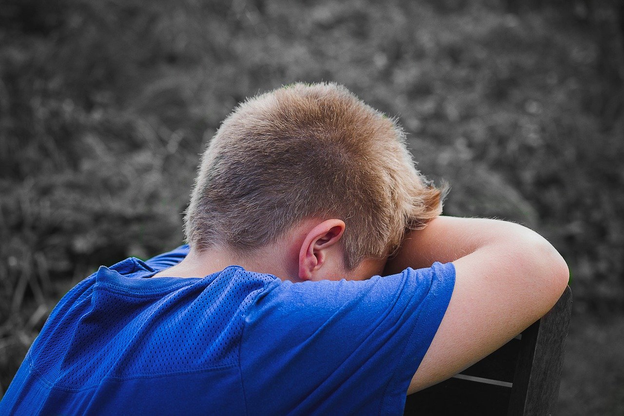 Ein kleiner Junge mit kurzen blonden Haaren und blauem Hemd lehnt seinen Kopf auf verschränkten Armen, mit dem Rücken zur Kamera, und wirkt verärgert oder nachdenklich. Der unscharfe Außenhintergrund lässt vermuten, dass er sich in der Nähe der Hochschule Coburg befindet und in Gedanken über seine Zukunft versunken ist.