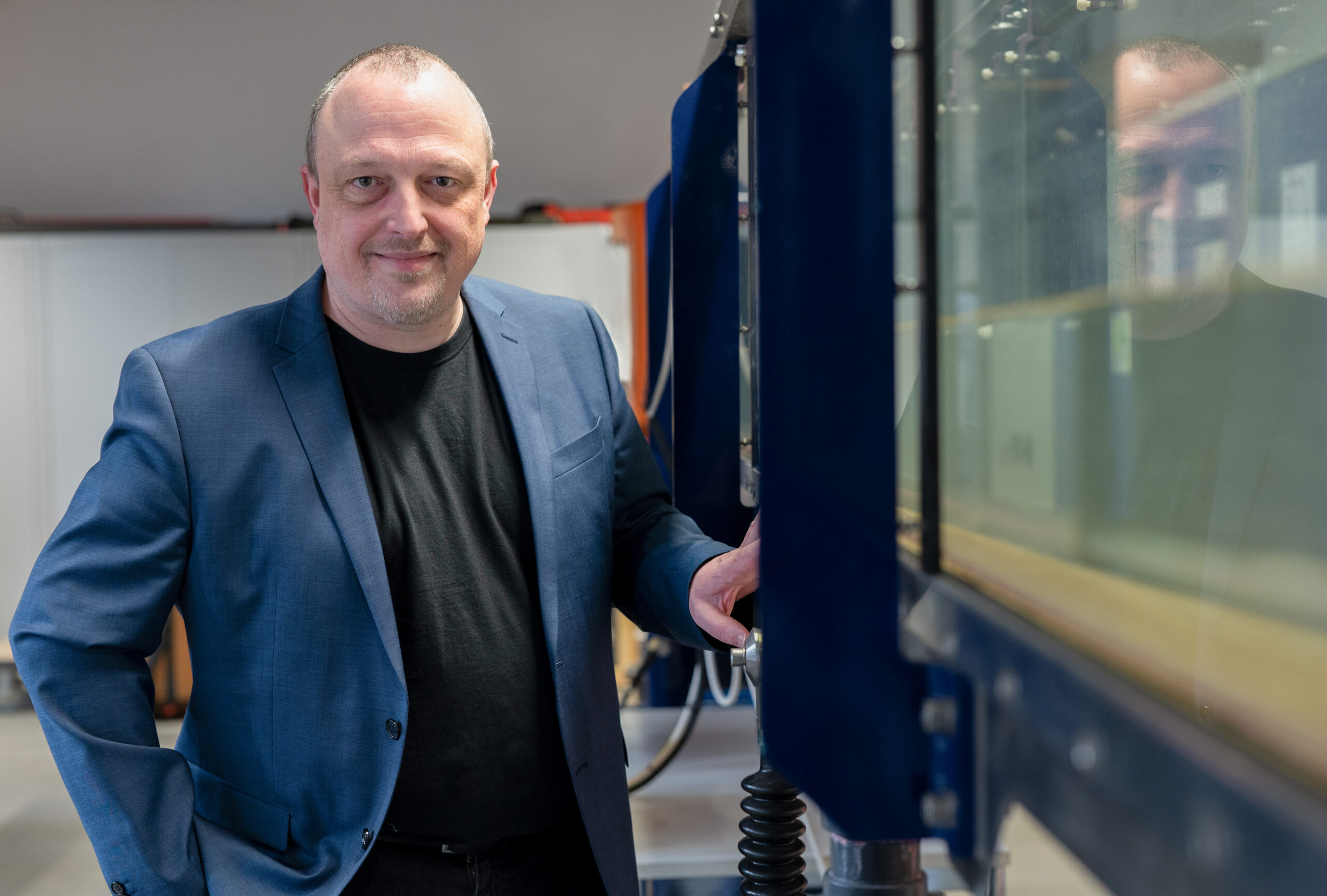 Ein Mann in blauem Blazer und schwarzem Hemd, der den Geist der Hochschule Coburg verkörpert, steht mit einem leichten Lächeln neben Industrieanlagen. Im Hintergrund ist ein verschwommener Innenraum voller Regale und Maschinen zu sehen.