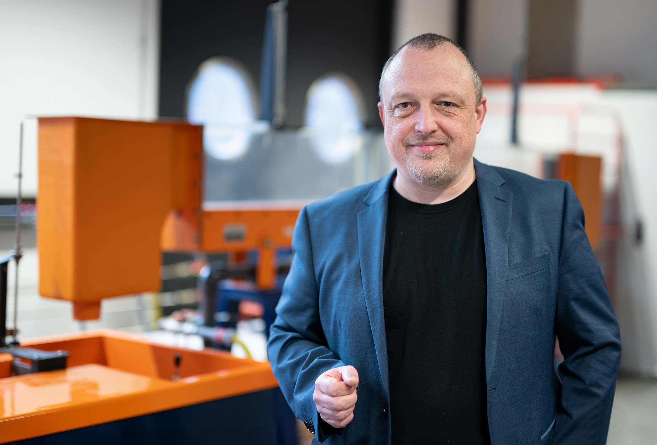 Ein Mann in blauem Blazer und schwarzem Hemd steht in einer Industrieanlage der Hochschule Coburg und lächelt in die Kamera. Er steht vor leuchtend orangefarbenen Maschinen, wobei Fenster und verschiedene Geräte die Hintergrundszenerie subtil aufwerten.