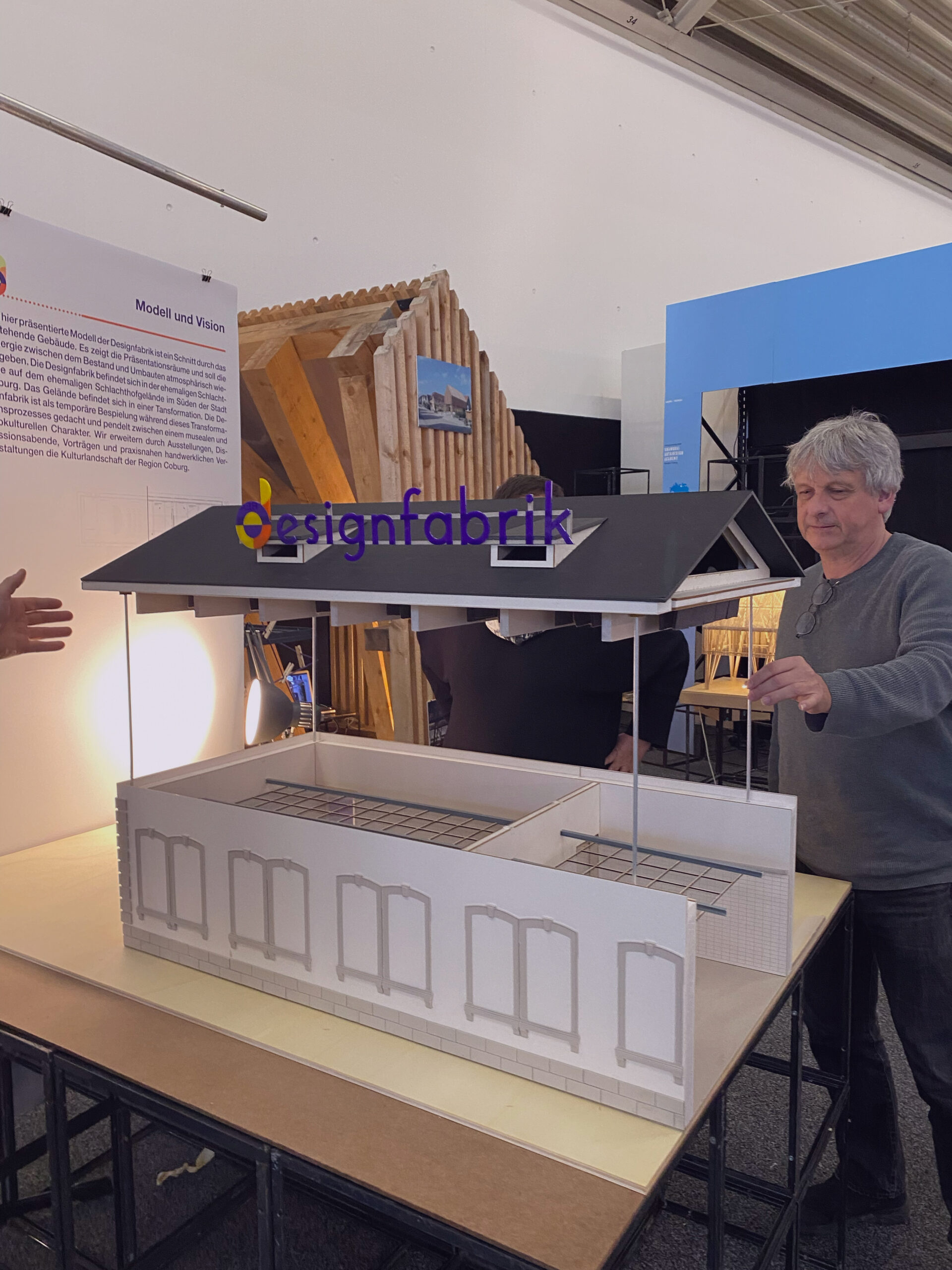 Zwei Personen stehen um ein detailreiches Architekturmodell eines Hauses mit schwarzem Dach und offenen Wänden herum. Die Kulisse ist vermutlich eine Ausstellung oder ein Workshop an der Hochschule Coburg. Über ihnen prangt prominent das Wort „designfabrik“, das die kreative Zusammenarbeit in Aktion unterstreicht.