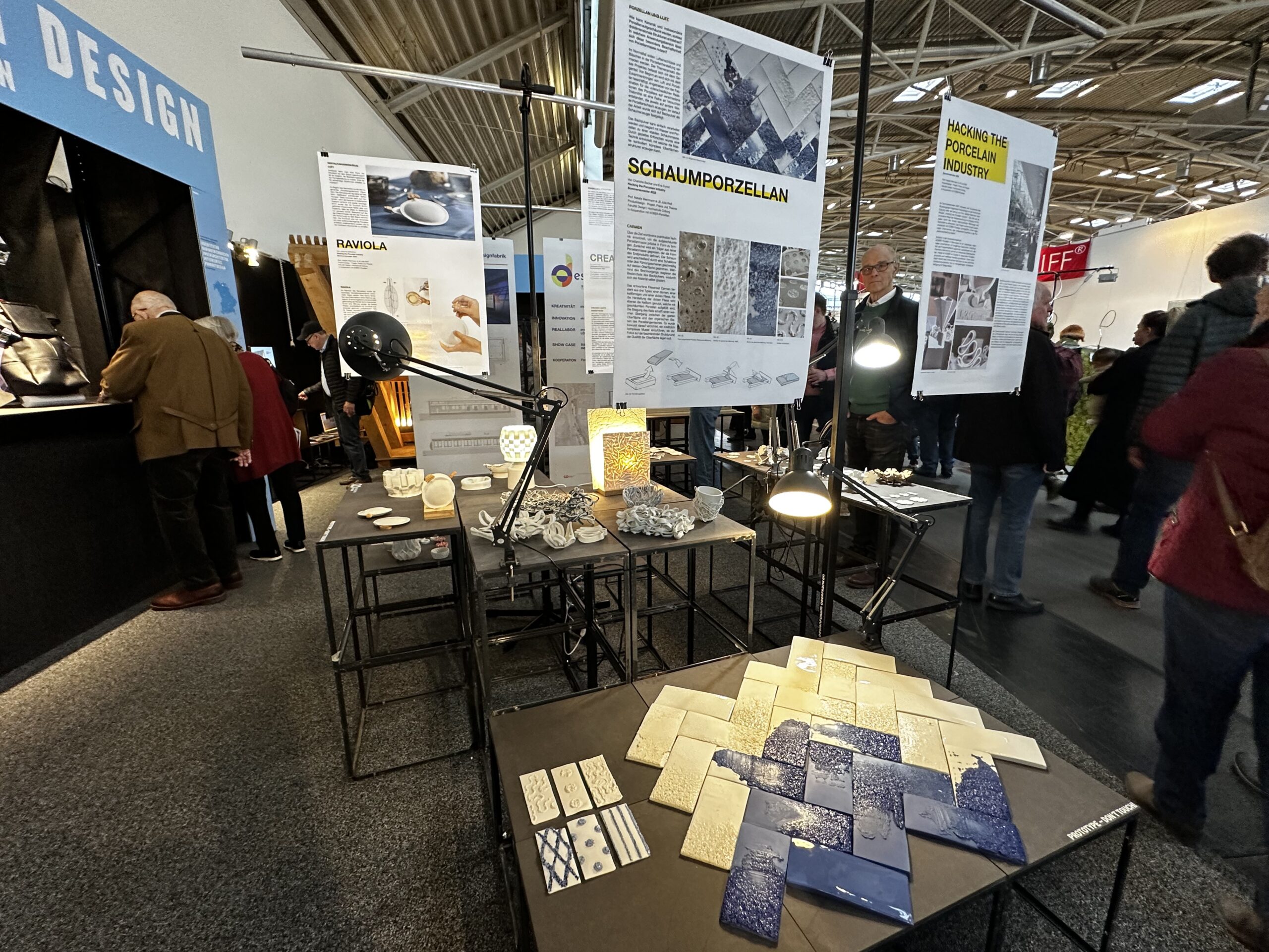 Am Design-Ausstellungsstand der Hochschule Coburg werden Keramik- und Porzellanstücke ausgestellt. Über den Exponaten hängen Plakate mit Text und Bildern, während die Besucher umhergehen und sie betrachten können. Die Beleuchtung ist hell und hebt die komplizierten Designs auf jedem Objekt hervor.