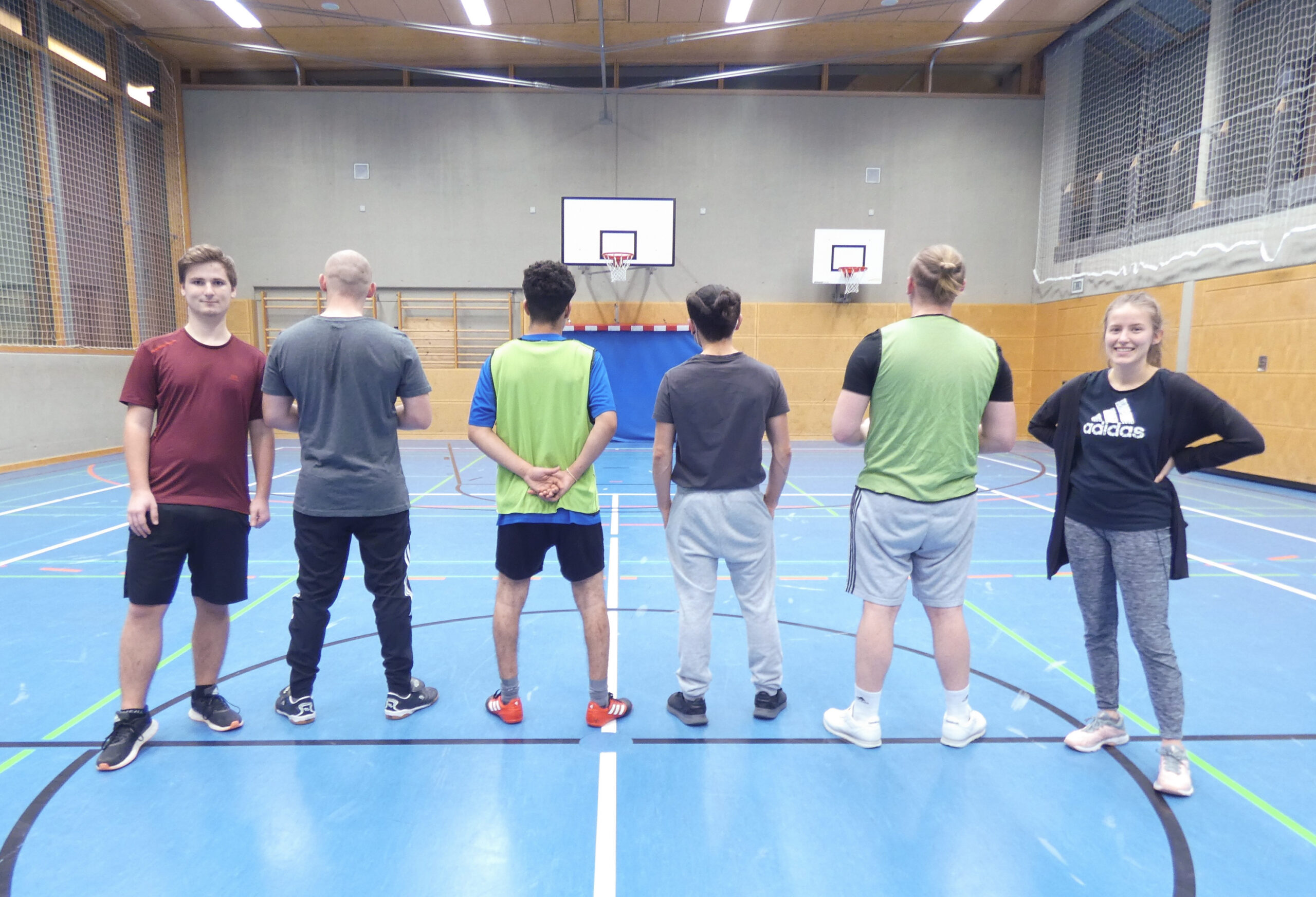 Sechs Personen stehen in der Turnhalle der Hochschule Coburg. Einer blickt nach vorne, die anderen nach hinten auf den blauen Basketballplatz. Die Szene mit den Holzwänden und Basketballkörben zeigt sie in ihrer Sportkleidung, bereit zum Einsatz.