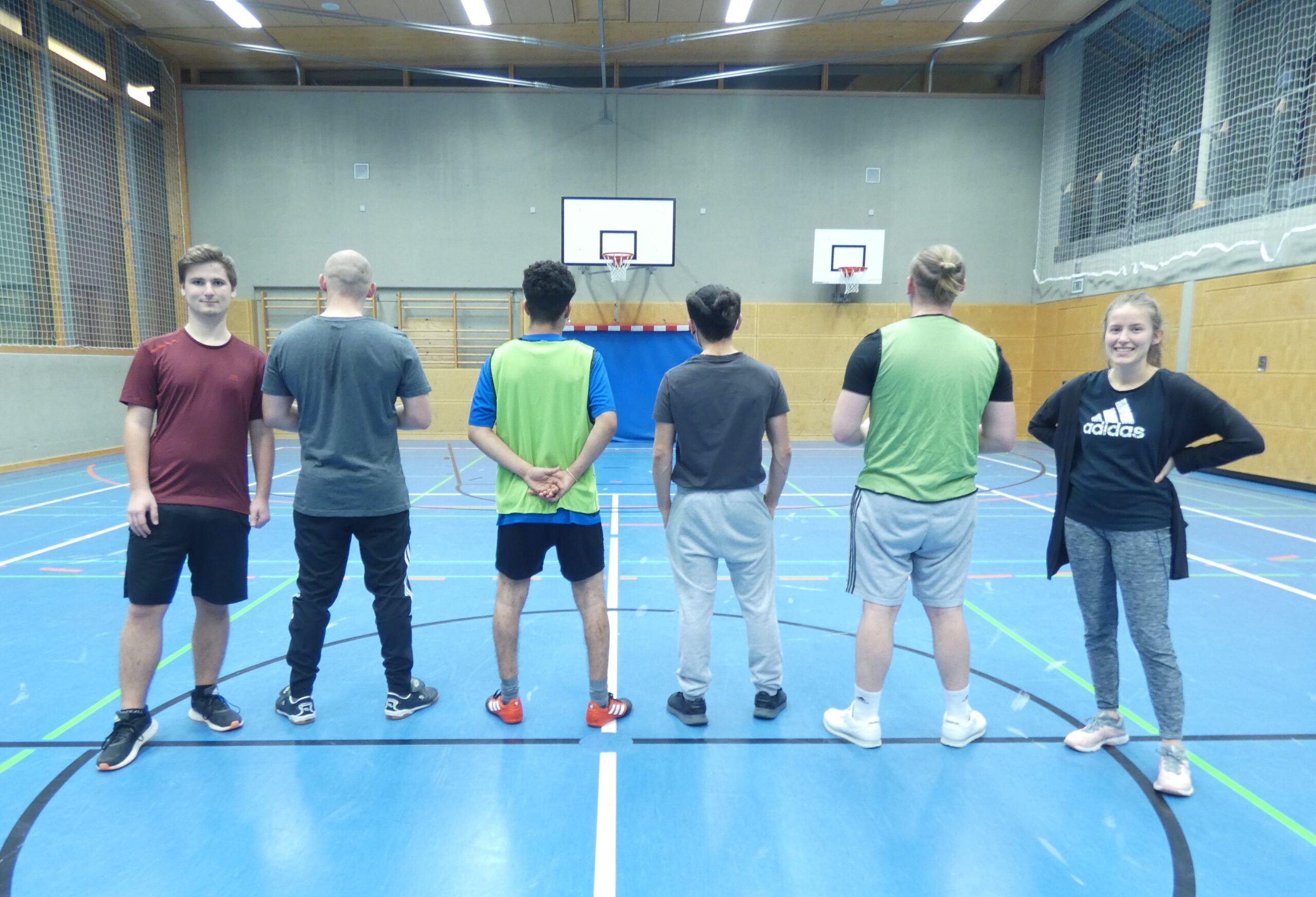 Fünf Personen stehen in einer Turnhalle der Hochschule Coburg vor einem Basketballkorb. Vier tragen Sportkleidung und kehren der Kamera den Rücken zu, während einer rechts nach vorne blickt und selbstbewusst lächelt und die Arme verschränkt. Die Turnhalle ist mit blauem Boden und Holzpaneelen ausgestattet und spiegelt den lebendigen Geist der Coburger Gemeinschaft wider.