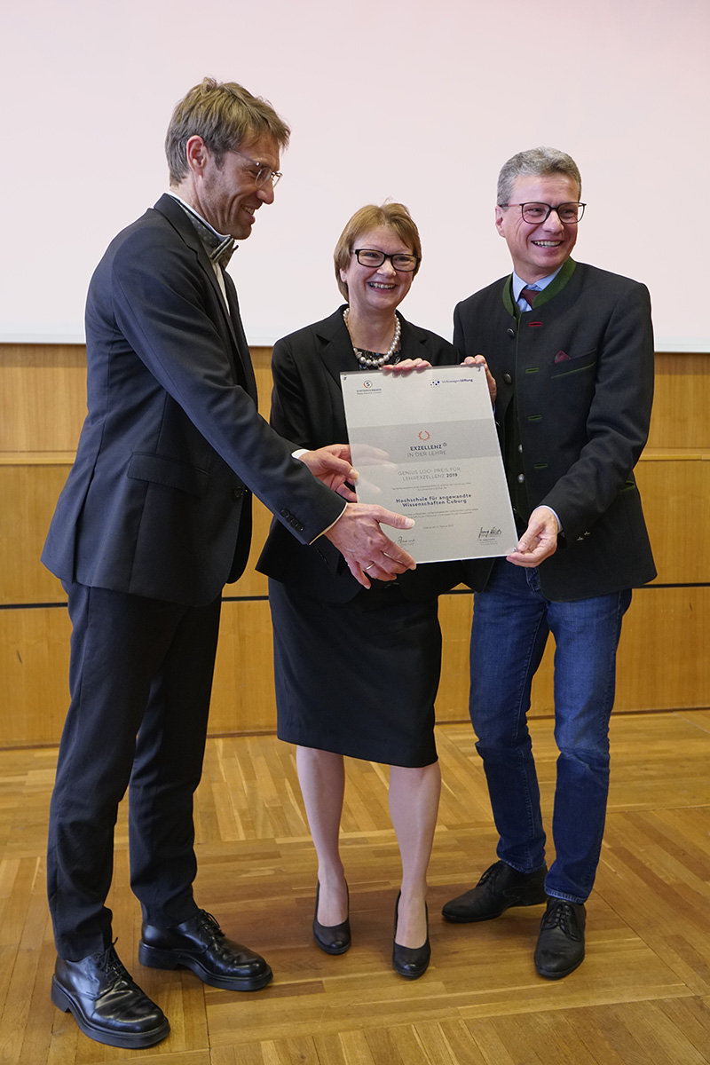 Drei Personen stehen lächelnd nebeneinander und halten stolz eine große Urkunde der Hochschule Coburg in den Händen. Sie befinden sich in einem Innenraum mit Holzboden und einer hellen Wand. Die Frau in der Mitte trägt ein schwarzes Outfit, während die beiden Männer Anzüge tragen.