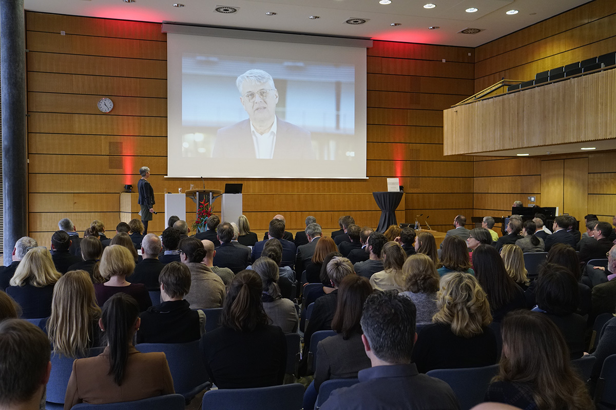 Im elegant holzgetäfelten Konferenzraum der Hochschule Coburg steht ein Redner an einem Podium und spricht zu einem sitzenden Publikum. Auf einem großen Bildschirm hinter dem Redner wird eine Person gezeigt, hervorgehoben durch rote Lichtakzente, die der Veranstaltung Eleganz verleihen.