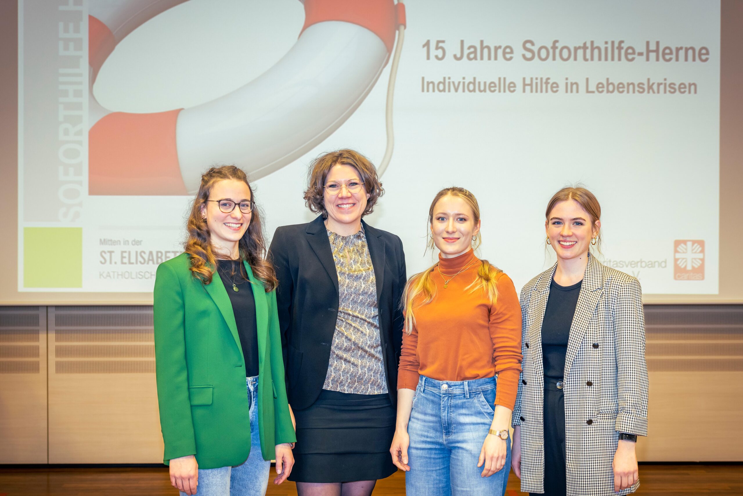 Vier Frauen stehen Seite an Seite und lächeln in legerer Business-Kleidung in die Kamera. Hinter ihnen informiert eine Präsentationswand über das 15-jährige Jubiläum eines Krisenhilfe-Programms an der Hochschule Coburg.