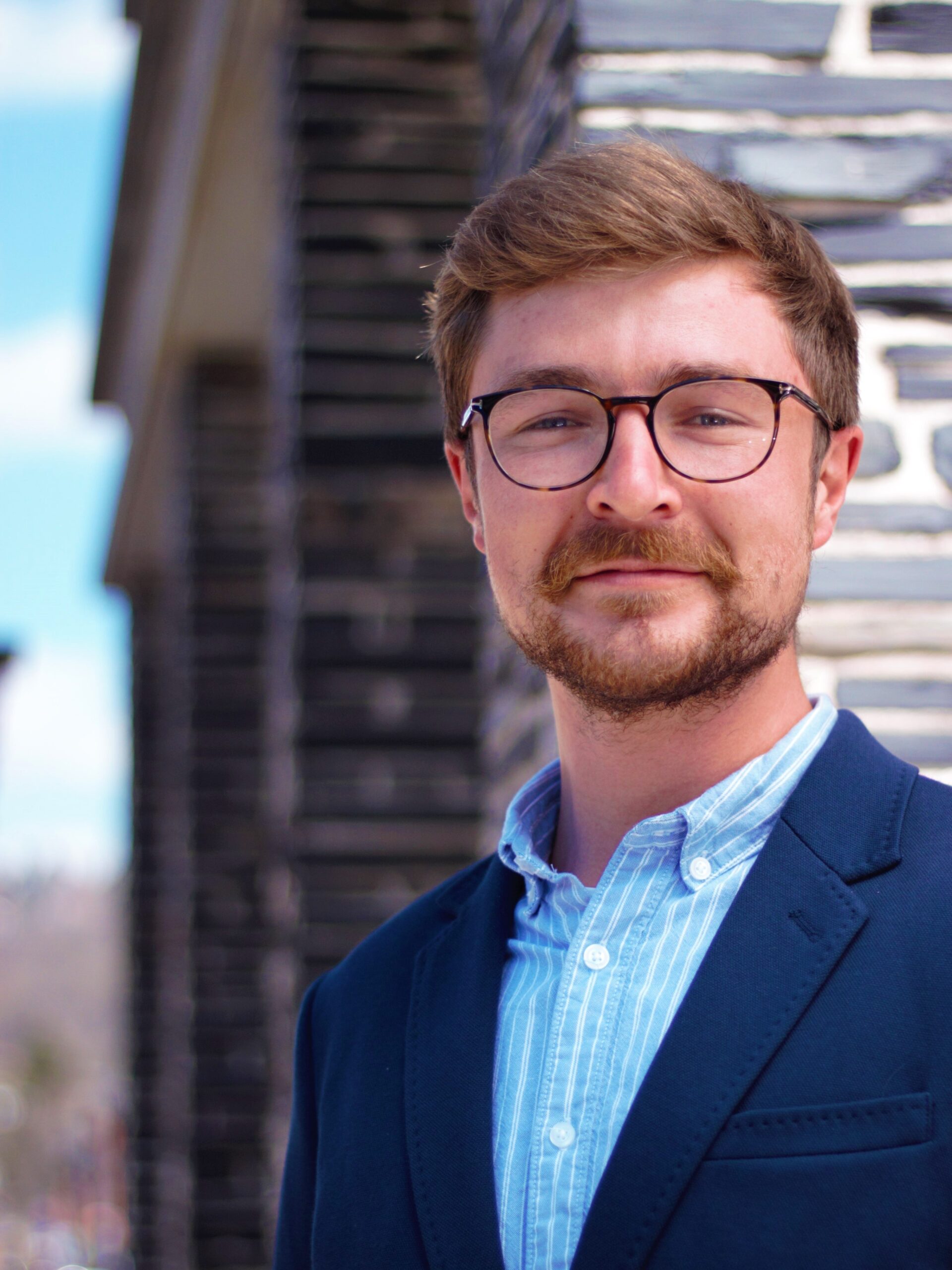 Ein Mann mit Brille, Schnurrbart und kurzen Haaren trägt einen blauen Blazer über einem Hemd der Hochschule Coburg. Er steht neben einer Steinmauer und wird von sanftem Tageslicht erhellt. Der Hintergrund ist leicht verschwommen.