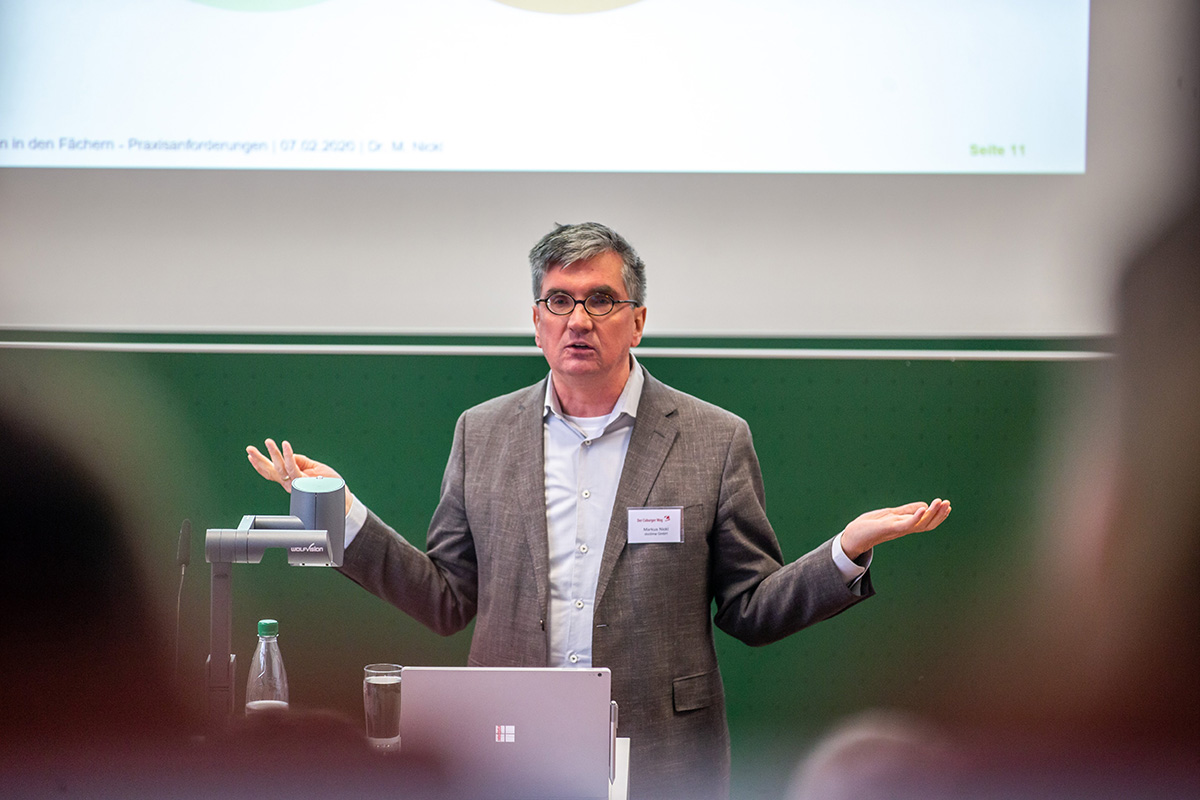 Ein Mann im Anzug gestikuliert mit beiden Händen, während er an einem mit einem Laptop und einer Flasche Wasser ausgestatteten Podium vor der grünen Kulisse der Hochschule Coburg spricht. Über ihm werden auf einer Projektionsfläche Text und visuelle Inhalte angezeigt, die seine Präsentation unterstreichen.