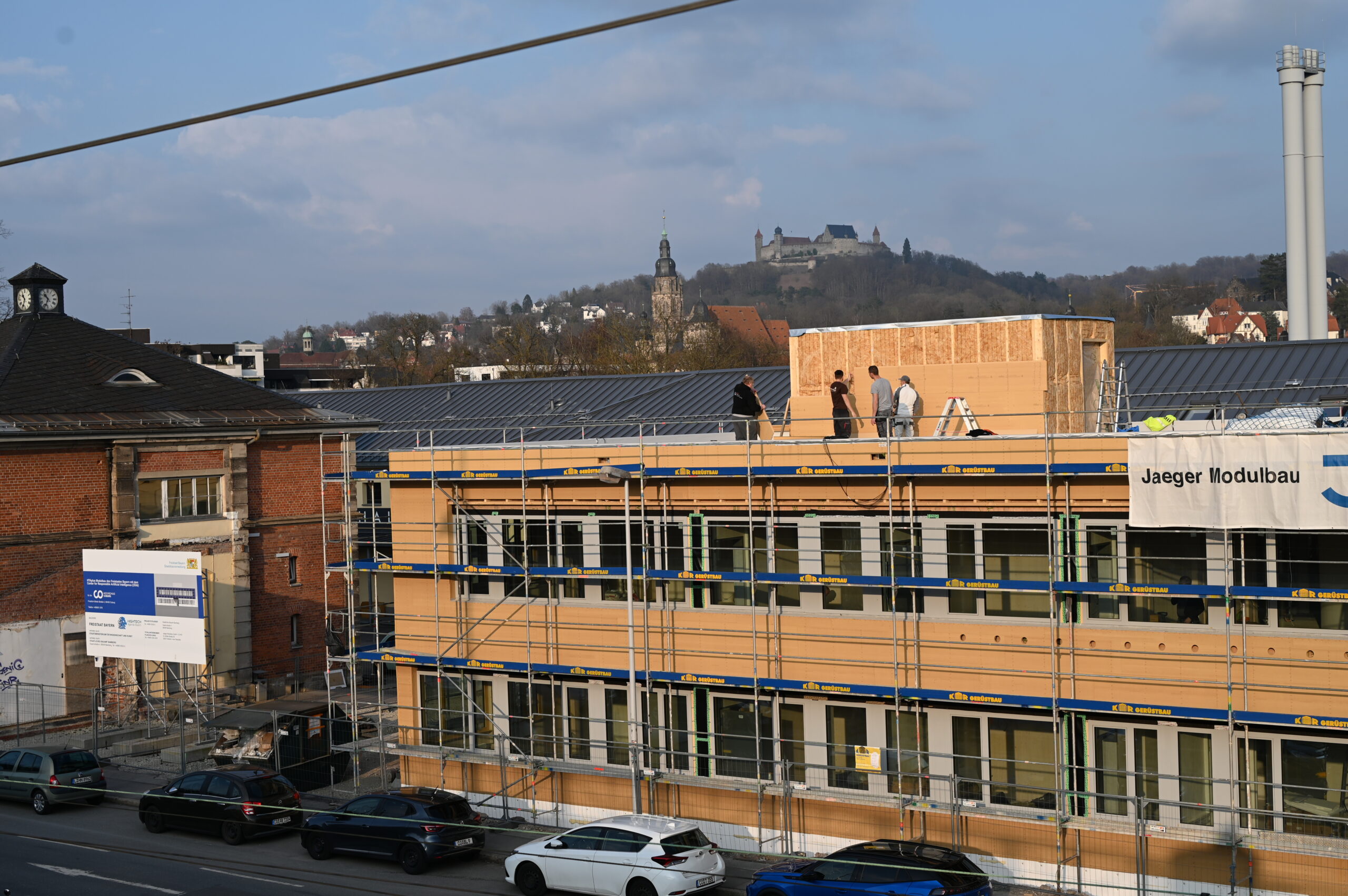 A construction site buzzes with activity as workers toil on the roof of a partially built modular building at Hochschule Coburg. Scaffolding encases the structure, while a hill crowned with a castle and church tower looms in the background. Cars line the street, framing this academic hub.