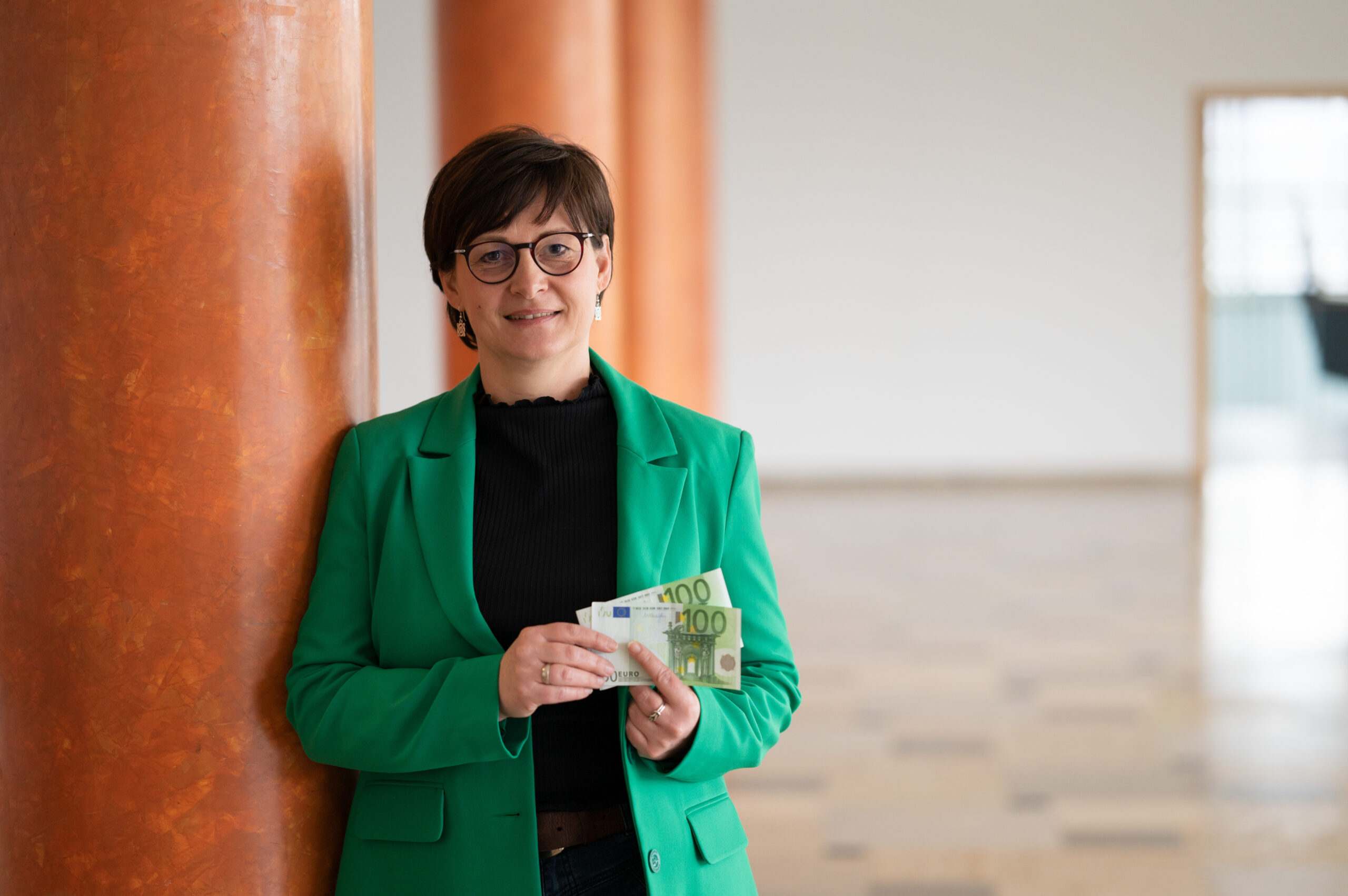 Eine Frau mit Brille und grünem Blazer, möglicherweise eine Fakultätsangehörige der Hochschule Coburg, steht in einem Innenbereich an einer orangefarbenen Säule und hält mehrere Euro-Banknoten in der Hand. Hinter ihr befindet sich ein geräumiger Bereich mit einer offenen Tür, durch die natürliches Licht in die Szene fällt.