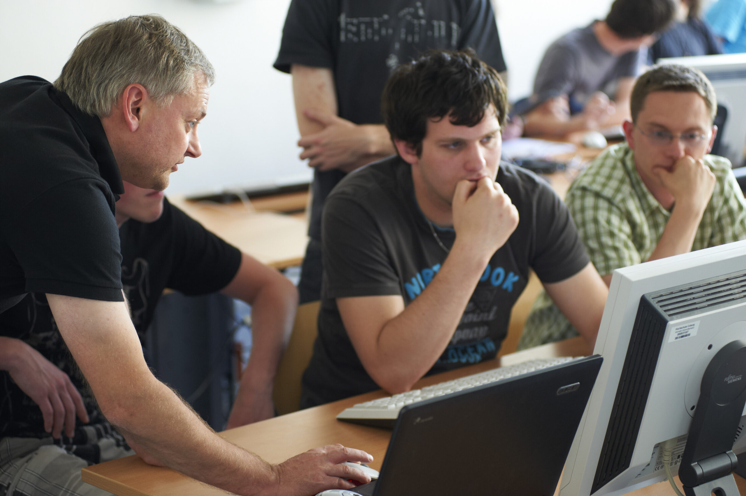 Eine Gruppe von vier Personen, drei Männer und eine Frau, sitzt an einem Schreibtisch in der Hochschule Coburg und schaut aufmerksam auf einen Computermonitor. Ein Mann zeigt auf den Bildschirm, während die anderen ihn genau beobachten, was den Eindruck erweckt, dass sie sich in einer Unterrichts- oder Büroumgebung befinden.