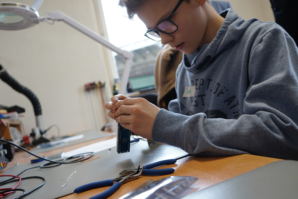 Ein junger Mensch mit Brille und Kapuzenpullover von der Hochschule Coburg arbeitet konzentriert an einer Platine, umgeben von elektronischen Bauteilen und Werkzeugen auf einem Tisch. Im Hintergrund sind eine Lupenlampe und andere Geräte zu sehen.