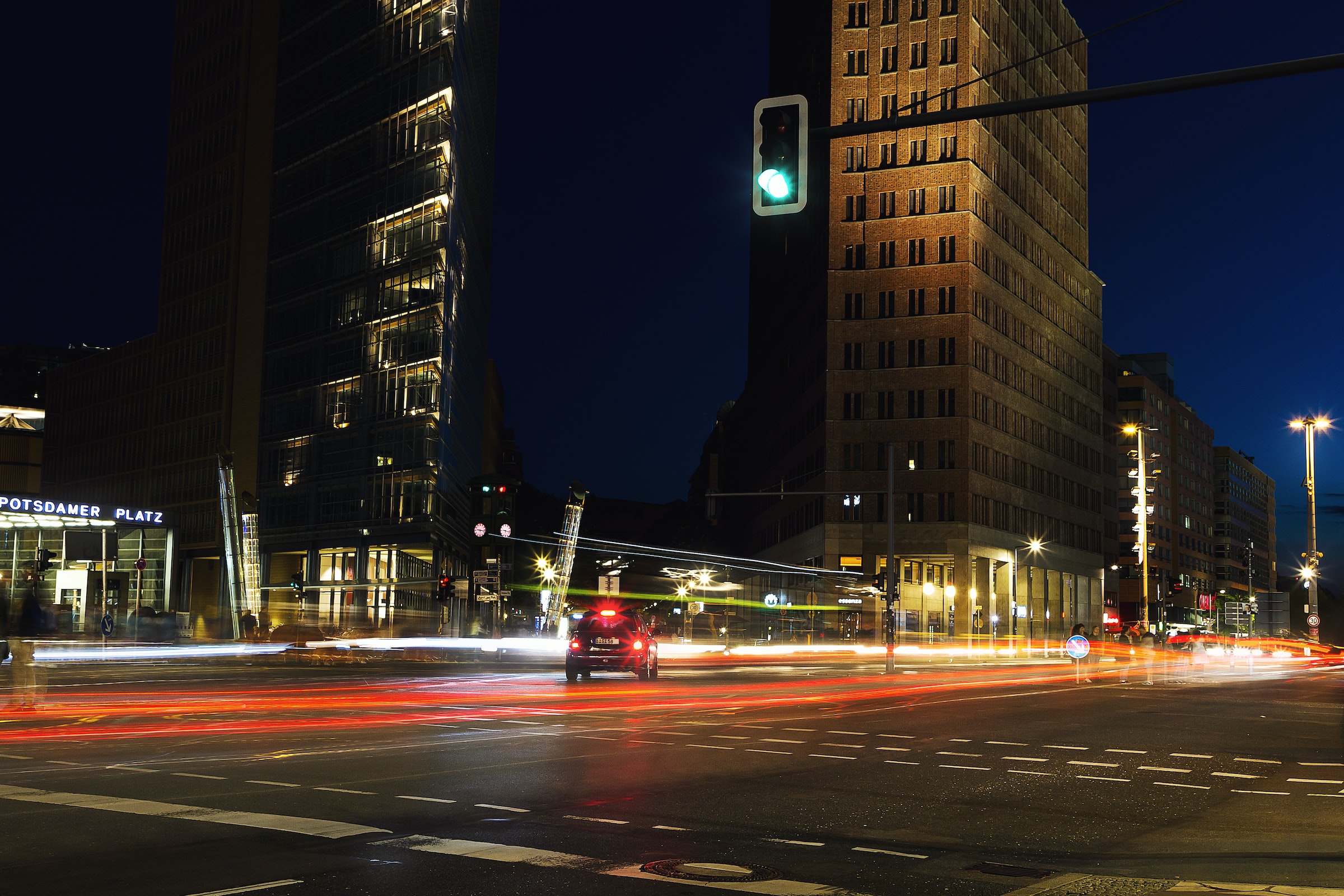 Nächtliche Stadtansicht am Potsdamer Platz in Berlin mit beleuchteten hohen Gebäuden, die an die an der Hochschule Coburg studierten Städtebaupläne erinnern. Eine grüne Ampel signalisiert über einer belebten Kreuzung mit Streifen roter und weißer Autolichter, die Bewegung und Aktivität einfangen.