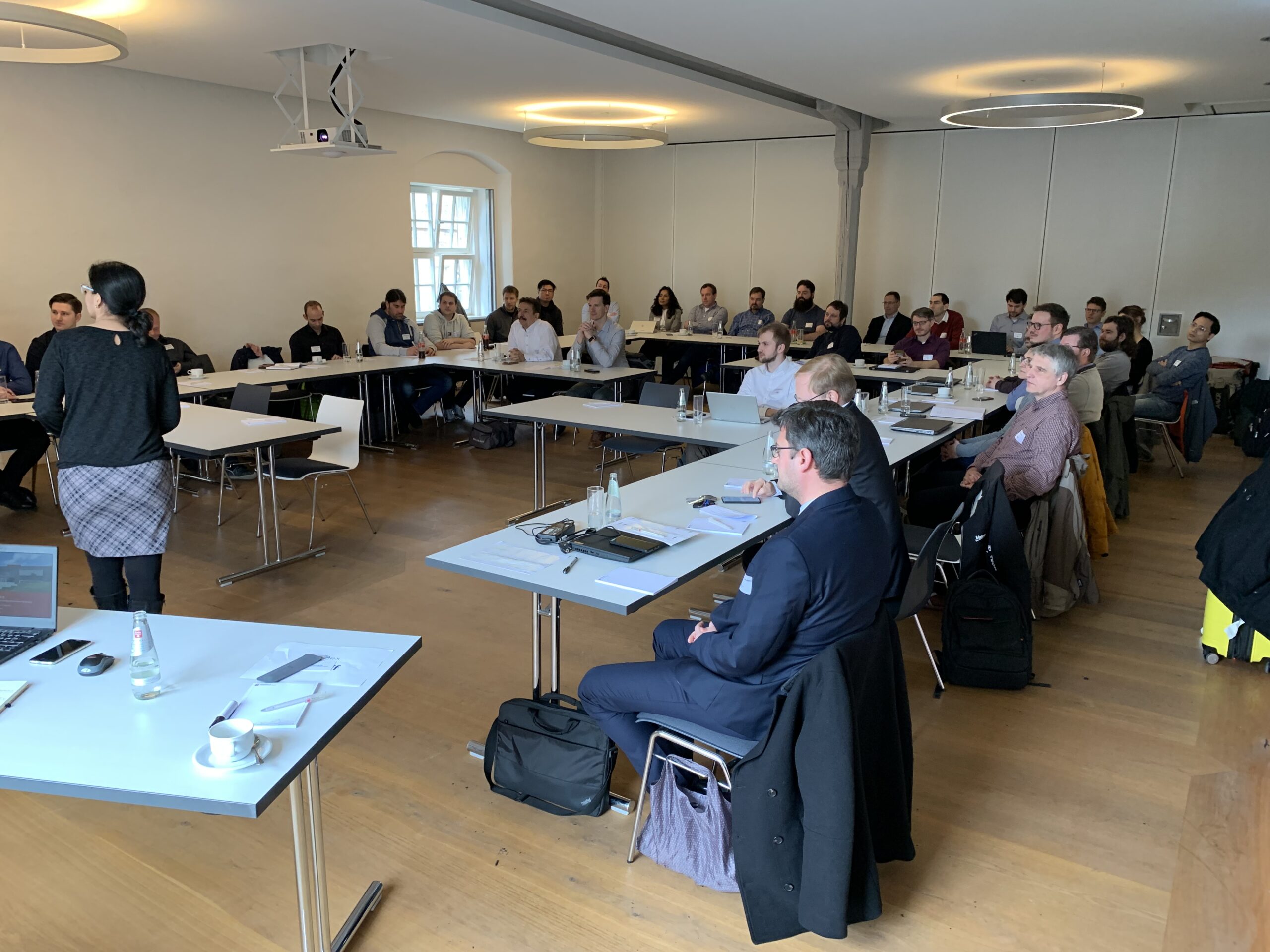 Eine Gruppe von Menschen sitzt in einem Konferenzraum der Hochschule Coburg, ganz vorne steht eine Frau, die vermutlich einen Vortrag hält. Die Tische sind U-förmig angeordnet, die Anwesenden hören aufmerksam zu.