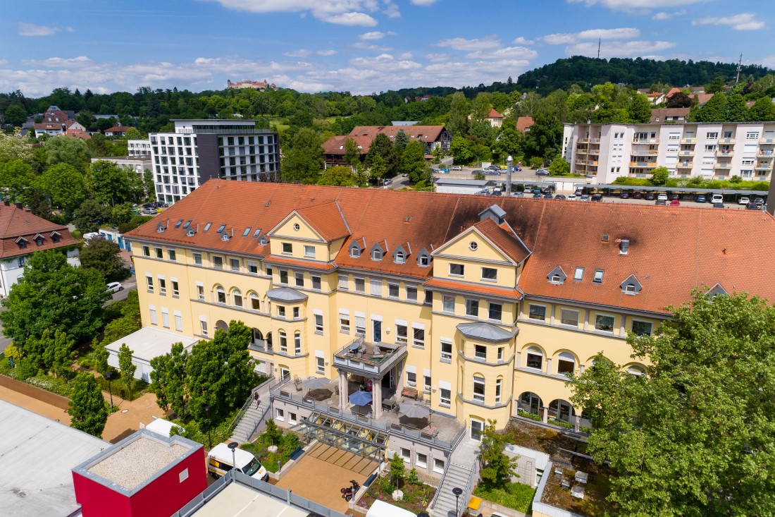 Ein großes, gelbes Gebäude mit rotem Ziegeldach, das an die Architektur der Hochschule Coburg erinnert, steht vor einer Kulisse aus üppigem Grün und hügeliger Landschaft. Umgeben von Bäumen und modernen Wohnhäusern liegt es unter einem blauen Himmel mit vereinzelten Wolken.