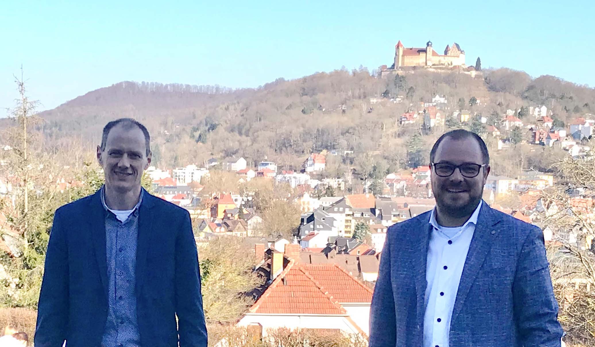 Zwei Männer in Anzügen stehen im Freien mit Blick auf eine Stadt und die Burg auf einem Hügel in der Nähe der Hochschule Coburg im Hintergrund. Der Himmel ist klar und blau, was die Sichtbarkeit der Landschaft erhöht.