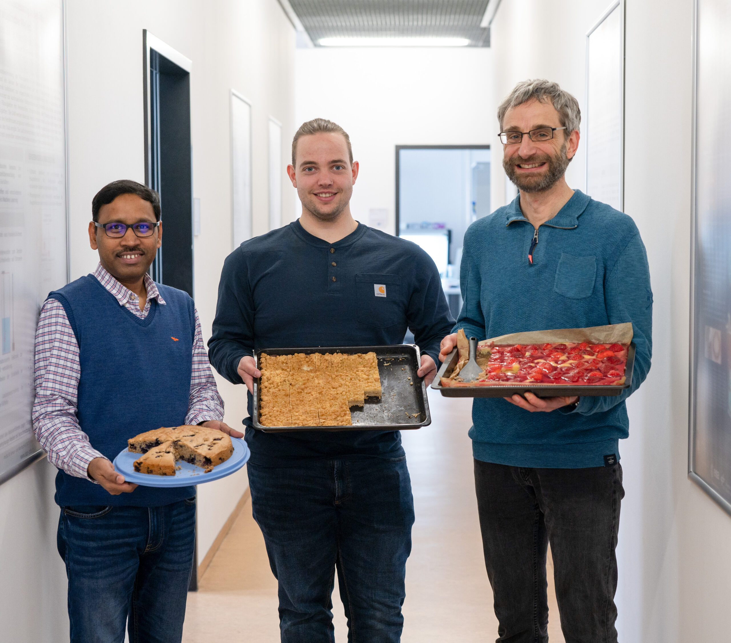 Three smiling men stand in a hallway at Hochschule Coburg, holding plates of baked goods: muffins, cookies, and a fruit-topped pastry. Dressed casually, they appear to be participating in a bake sale or potluck event.