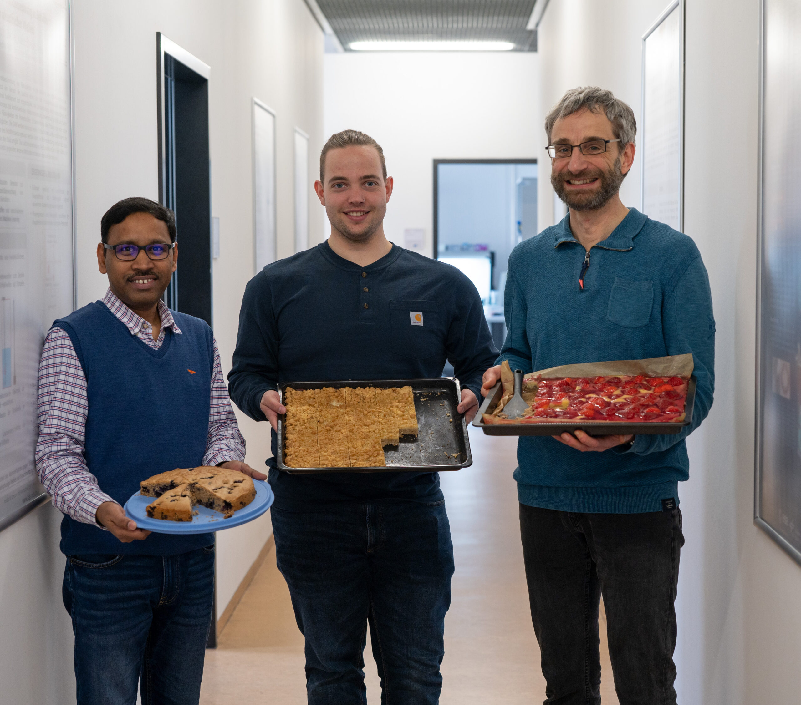 Three people stand in a hallway at Hochschule Coburg, each holding a tray of baked goods. The first person has cookies, the second a crumbly dessert, and the third a fruit-topped delight. They're all smiling warmly, dressed in casual clothes, embodying the spirit of community and sharing.