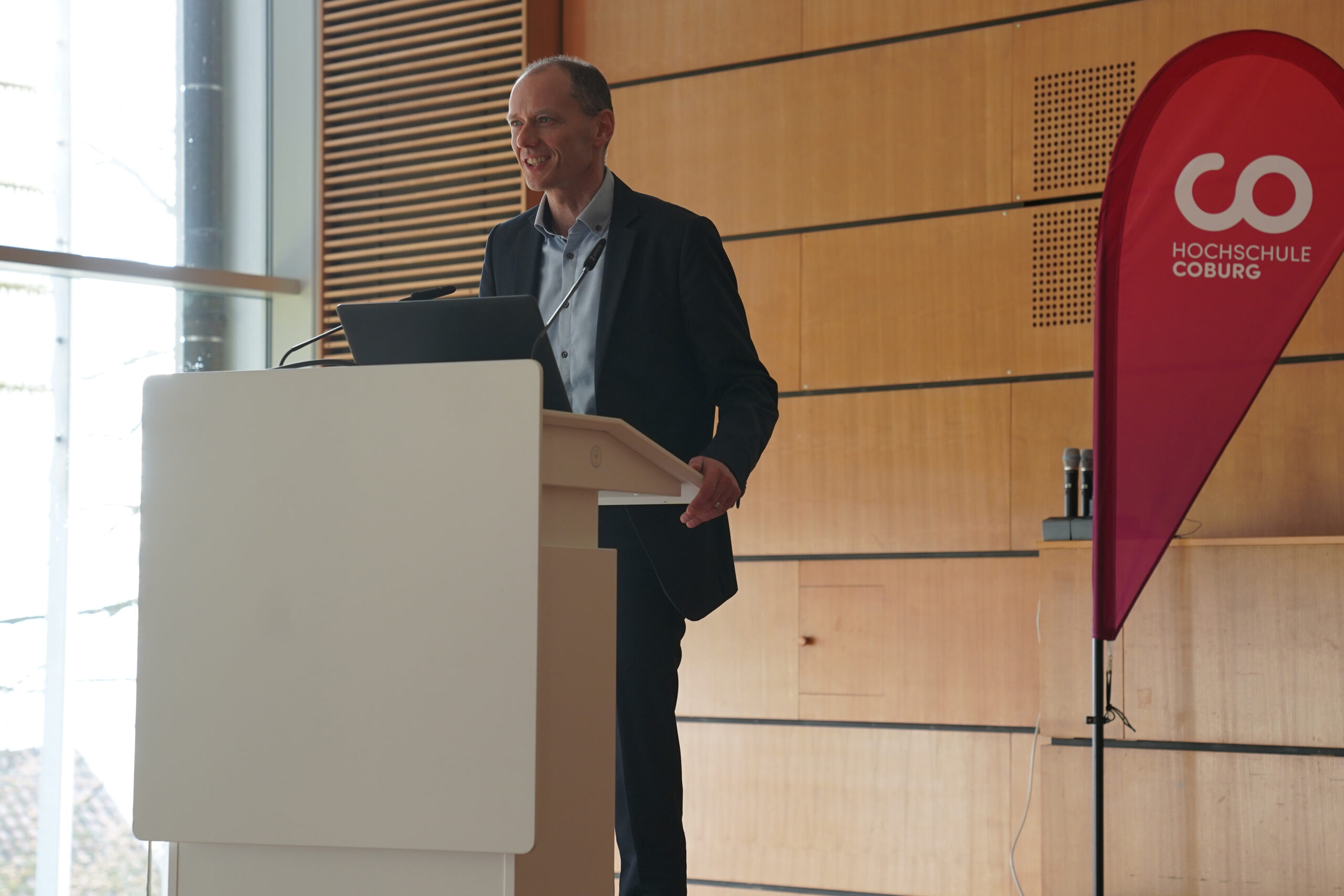 A person stands at a podium in a modern lecture hall, delivering a presentation. Dressed in a suit and smiling confidently, they have their laptop open beside them. Behind the speaker, a bold red banner proudly displays "Hochschule Coburg.