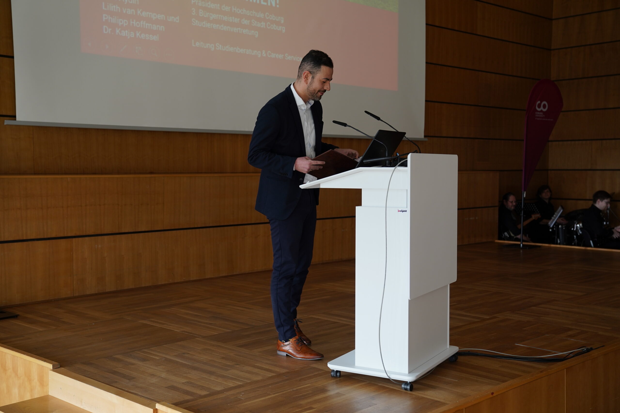 A man in a dark suit stands at a white podium, representing Hochschule Coburg, speaking into a microphone in a wooden-floored conference room. A large screen with text is projected behind him, capturing the attention of attendees seated in the background.