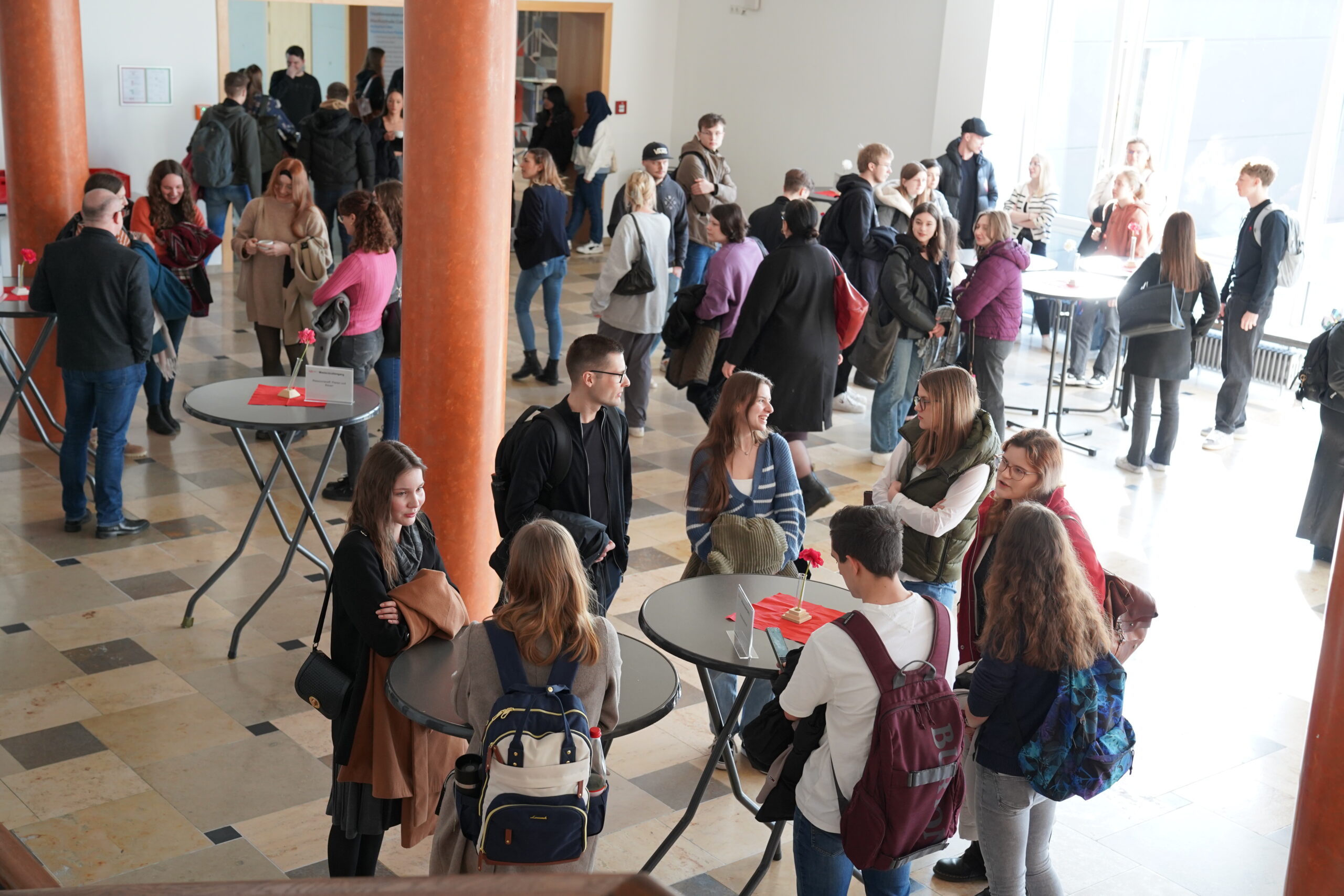 A group of people are gathered in a spacious indoor area at Hochschule Coburg with round tables. Some are chatting in small groups, while others stand alone. The setting appears bright and social, with a diverse mix of individuals engaged in conversation.