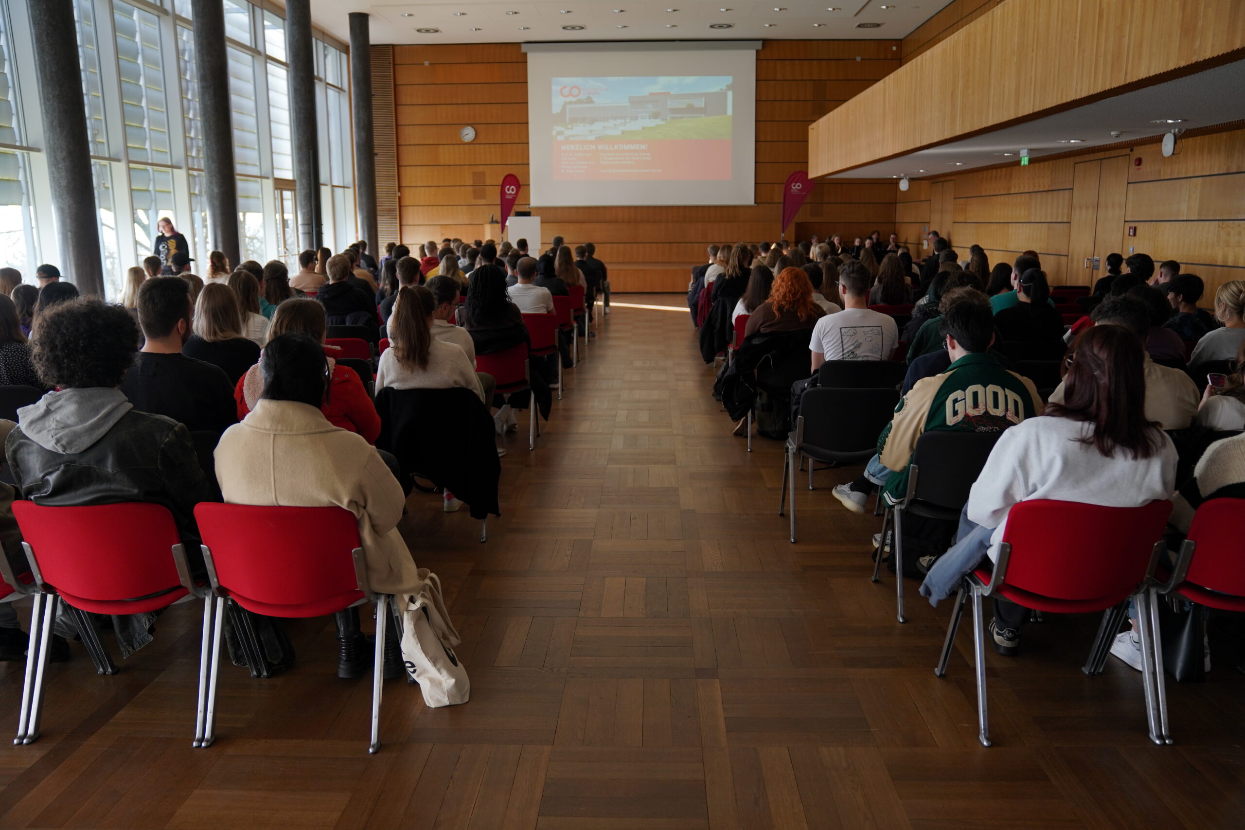 A large group of people is seated in a conference room at Hochschule Coburg, where wooden walls and flooring add warmth. They face a presentation screen displaying slides. The attentive audience enjoys natural light streaming in through tall windows on the left.