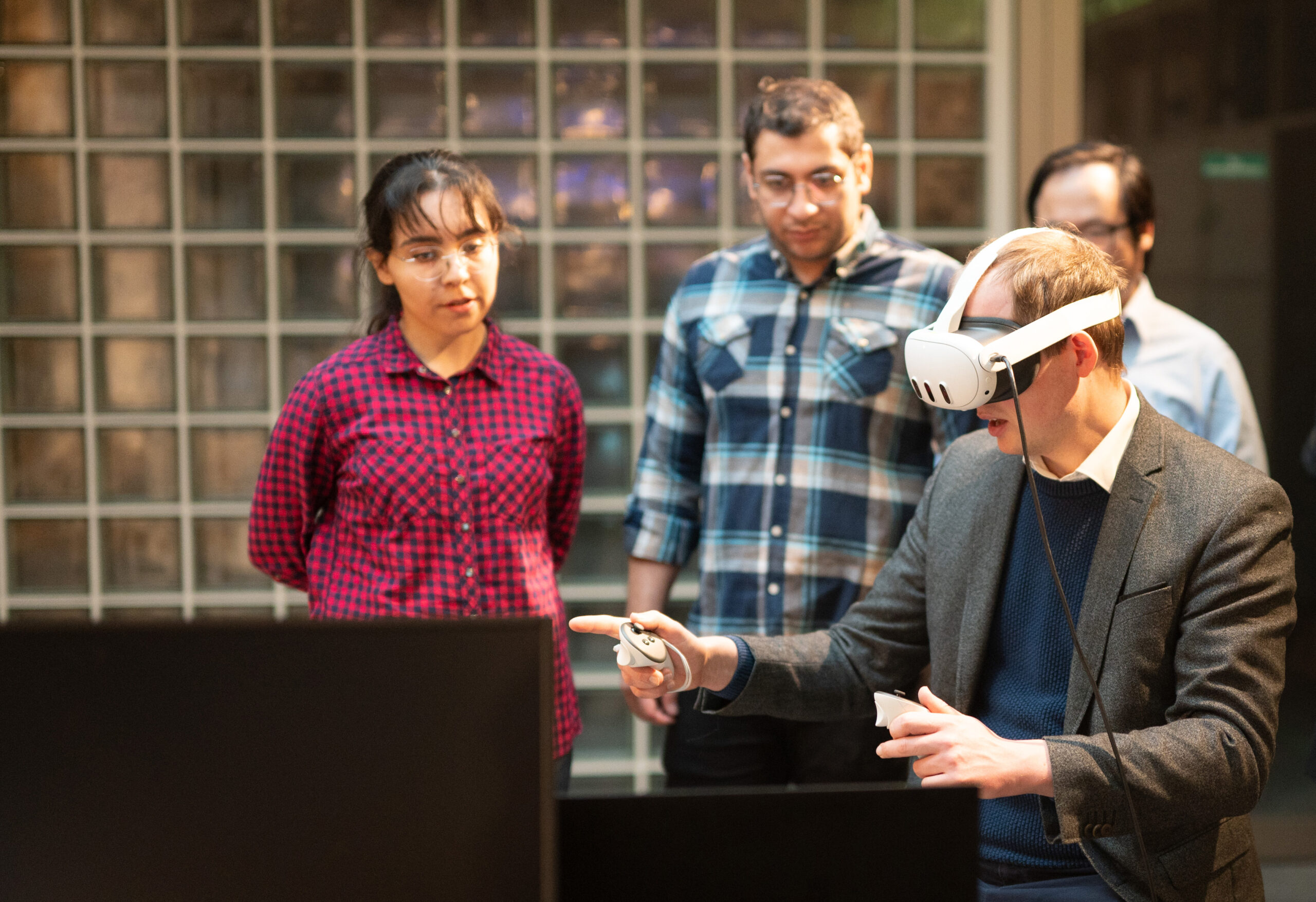 A man wearing a virtual reality headset and holding controllers interacts with a screen, capturing the attention of three onlookers. They stand in a room at Hochschule Coburg, framed by the modern touch of a glass block wall.