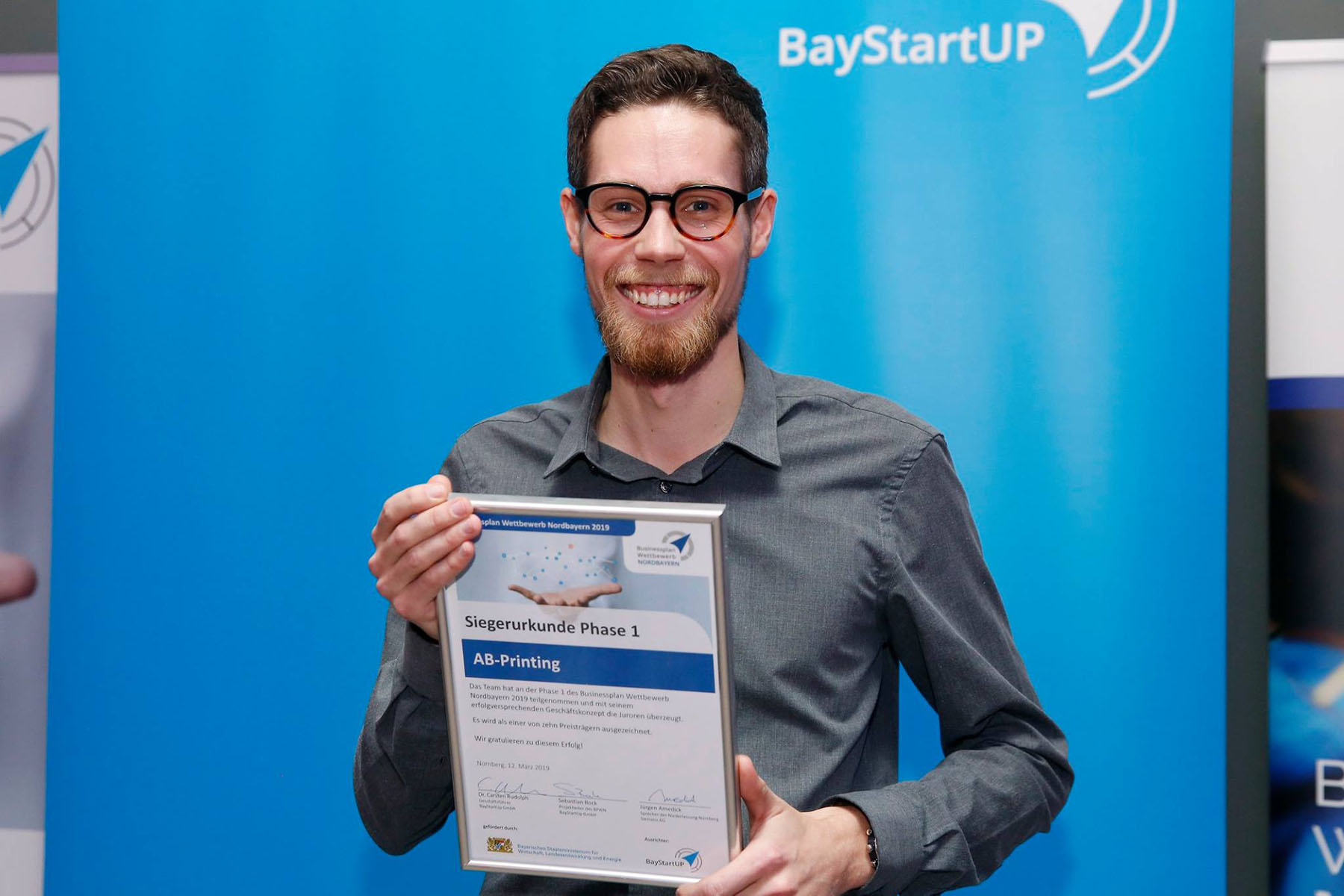 Eine Person mit Brille lächelt und hält stolz ein Zertifikat der Hochschule Coburg vor einem blauen BayStartUP-Banner.