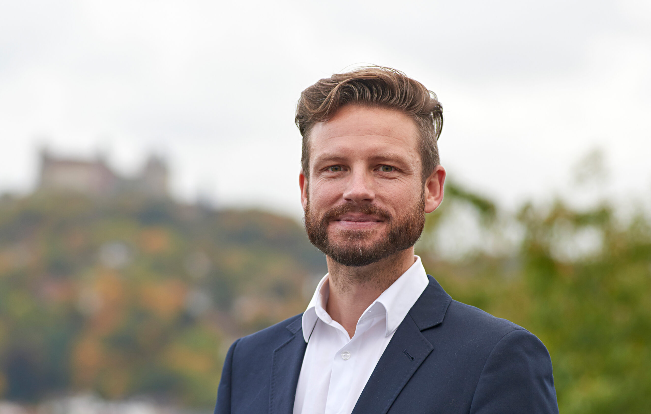 Ein Mann mit Vollbart und Schnurrbart steht im Freien auf dem Campus der Hochschule Coburg. Er trägt ein weißes Hemd und einen dunklen Blazer. Im Hintergrund ist eine verschwommene Landschaft mit Bäumen und einem entfernten Gebäude zu sehen. Er lächelt leicht und blickt in die Kamera.