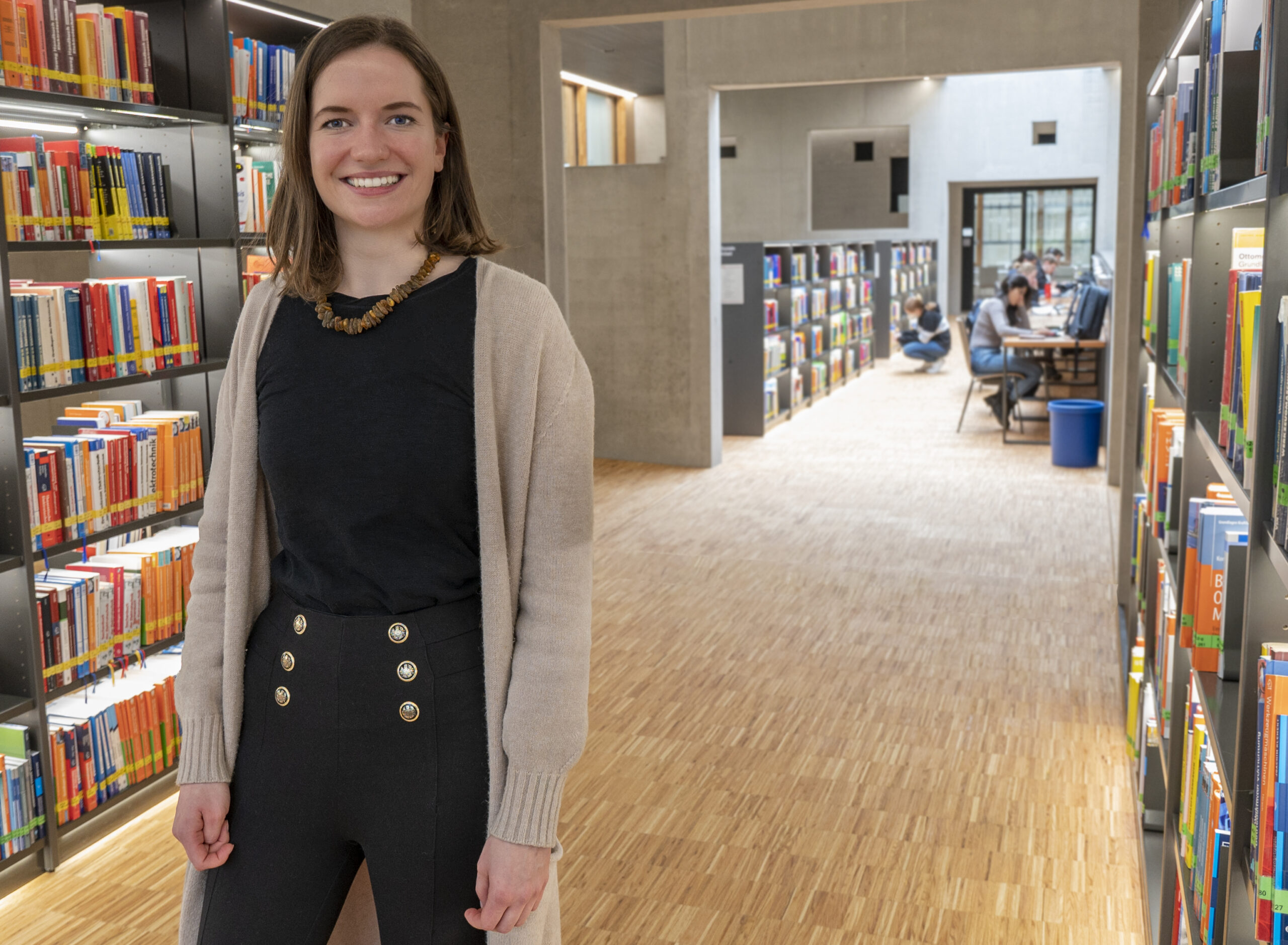 Eine Frau lächelt, während sie in der Bibliothek der Hochschule Coburg steht, umgeben von Bücherregalen voller bunter Bücher. Im Hintergrund lernen Menschen an einem Tisch. Boden und Wände sind aus hellem Holz, was eine warme Atmosphäre schafft.