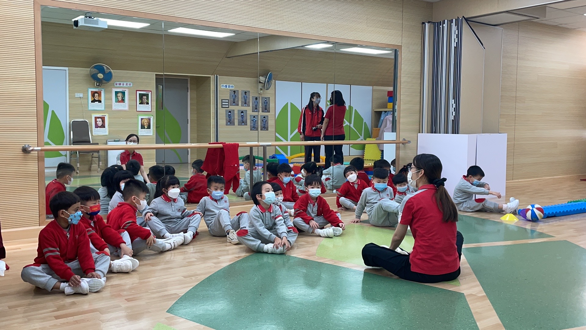 Eine Gruppe kleiner Kinder in passenden rot-grauen Uniformen sitzt auf dem polierten Holz- und grün gefliesten Boden der Turnhalle der Hochschule Coburg. Sie stehen einem Lehrer gegenüber, tragen alle Masken und sitzen neben einer Spiegelwand, die ihre aufmerksamen Ausdrücke widerspiegelt.