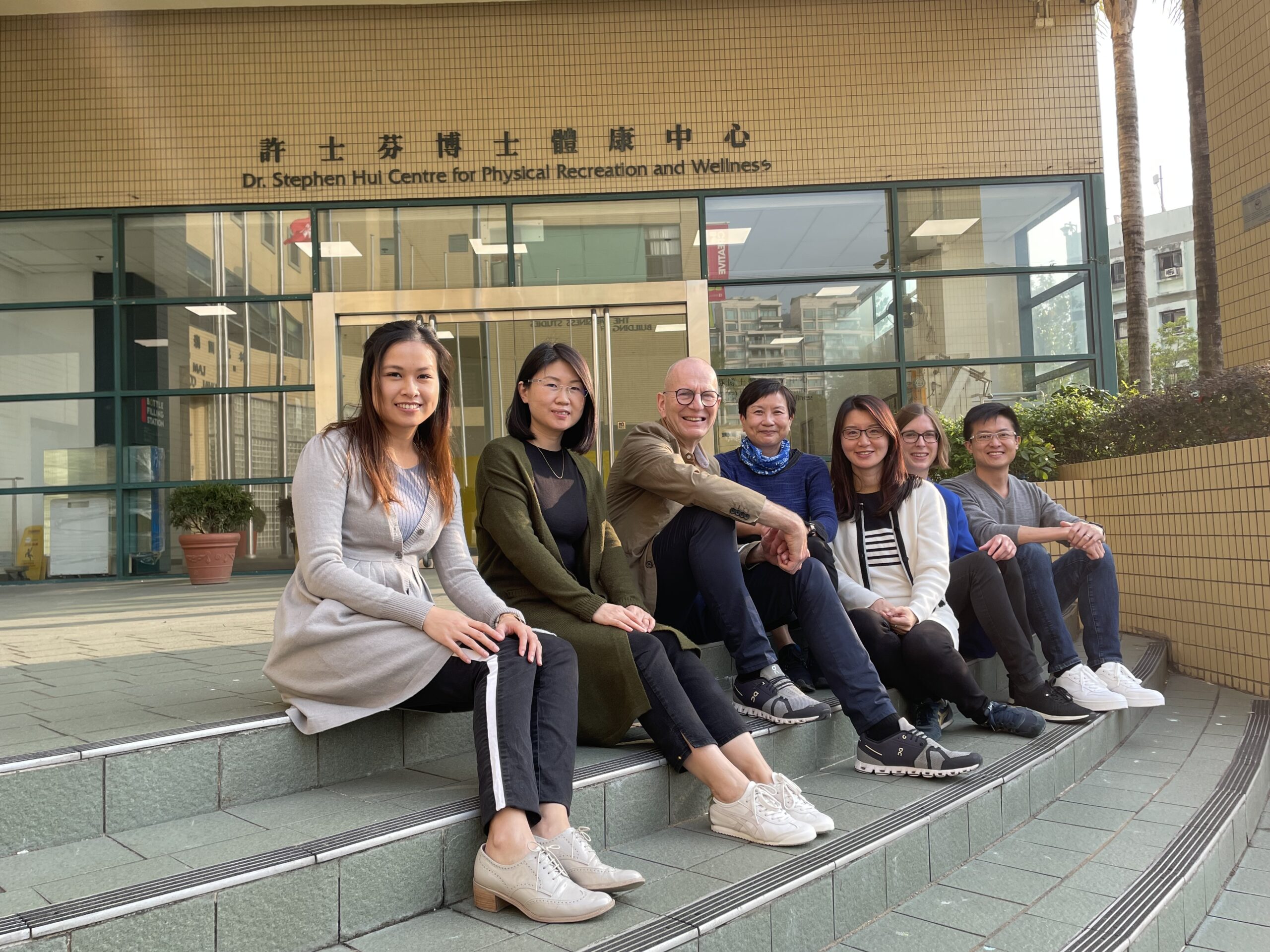 Eine Gruppe von sieben Personen sitzt auf den Stufen vor einem Gebäude mit der Aufschrift „Dr. Stephen Hui Centre for Physical Recreation and Wellness“ an der Hochschule Coburg. Sie lächeln und sind leger gekleidet, umgeben von Grünflächen und verkörpern das pulsierende Campusleben.