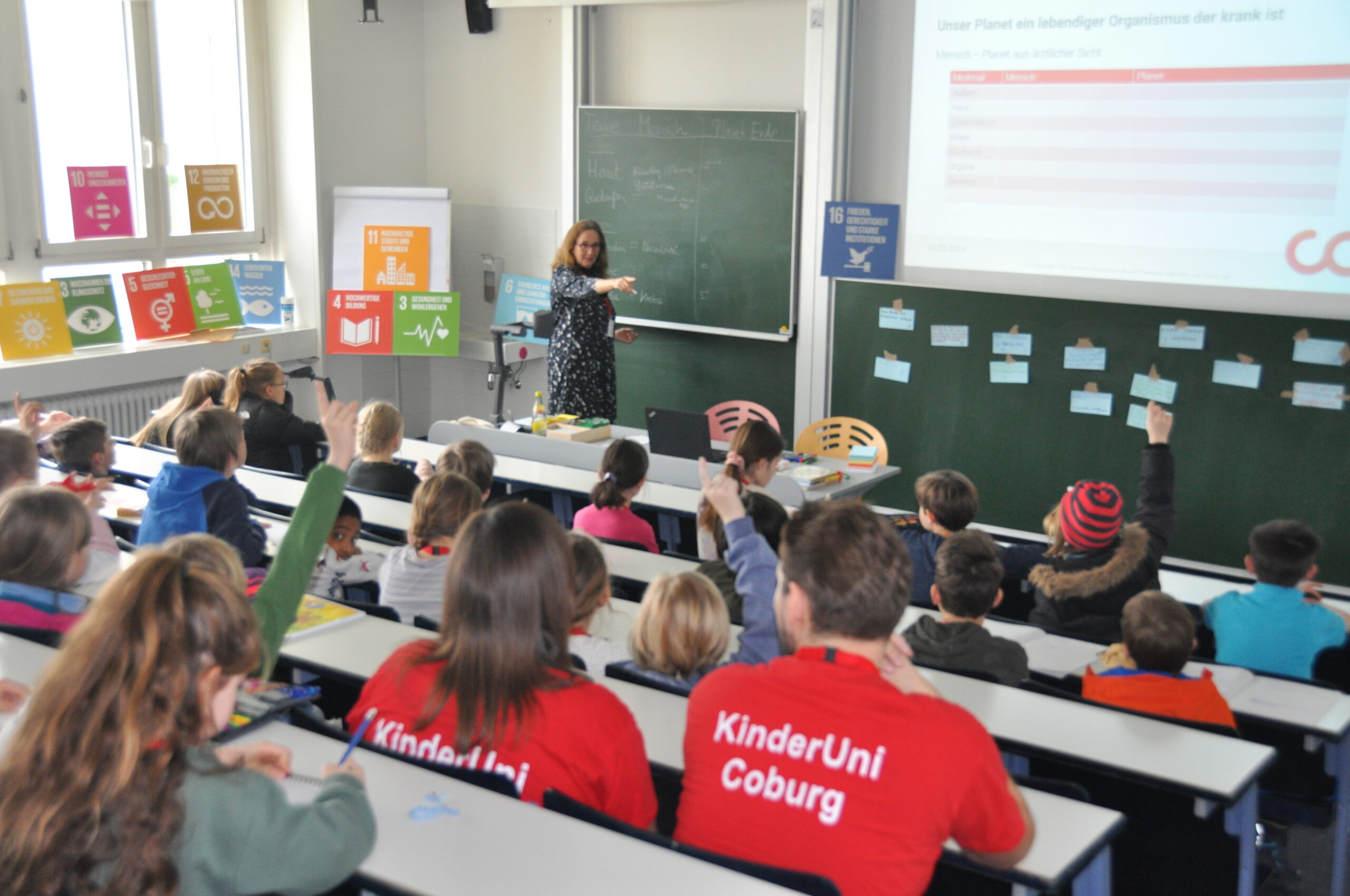 A woman stands at the front of a Hochschule Coburg classroom giving a presentation. Around twenty children are seated, some raising their hands. Posters about sustainability decorate the walls. The students wear red shirts with "KinderUni Coburg" printed on the back.