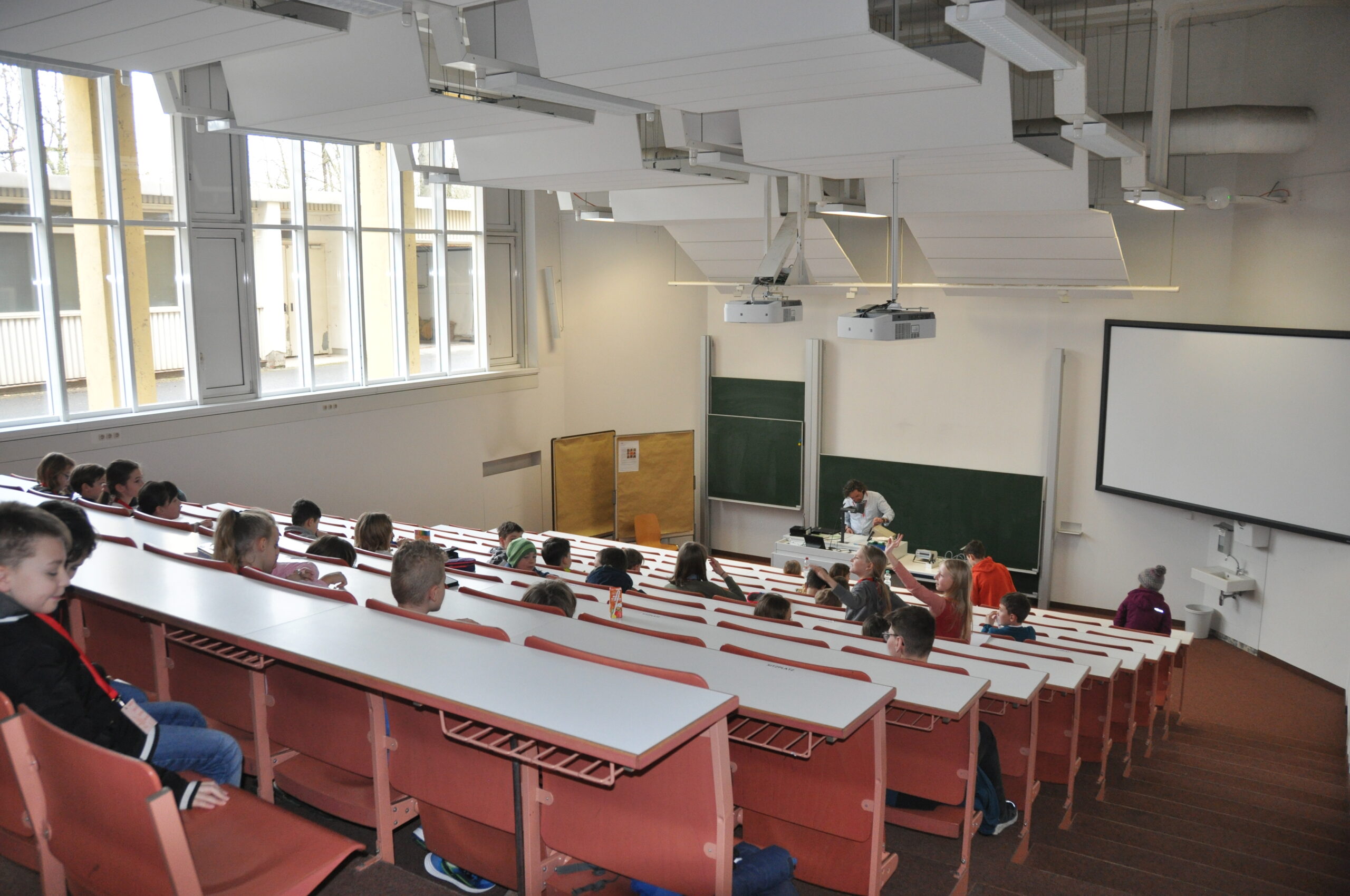 At the front of a large lecture hall in Hochschule Coburg, a professor addresses students seated at desks arranged in tiers. The room is spacious and well-lit, with two chalkboards and a projector screen prominently visible at the front.