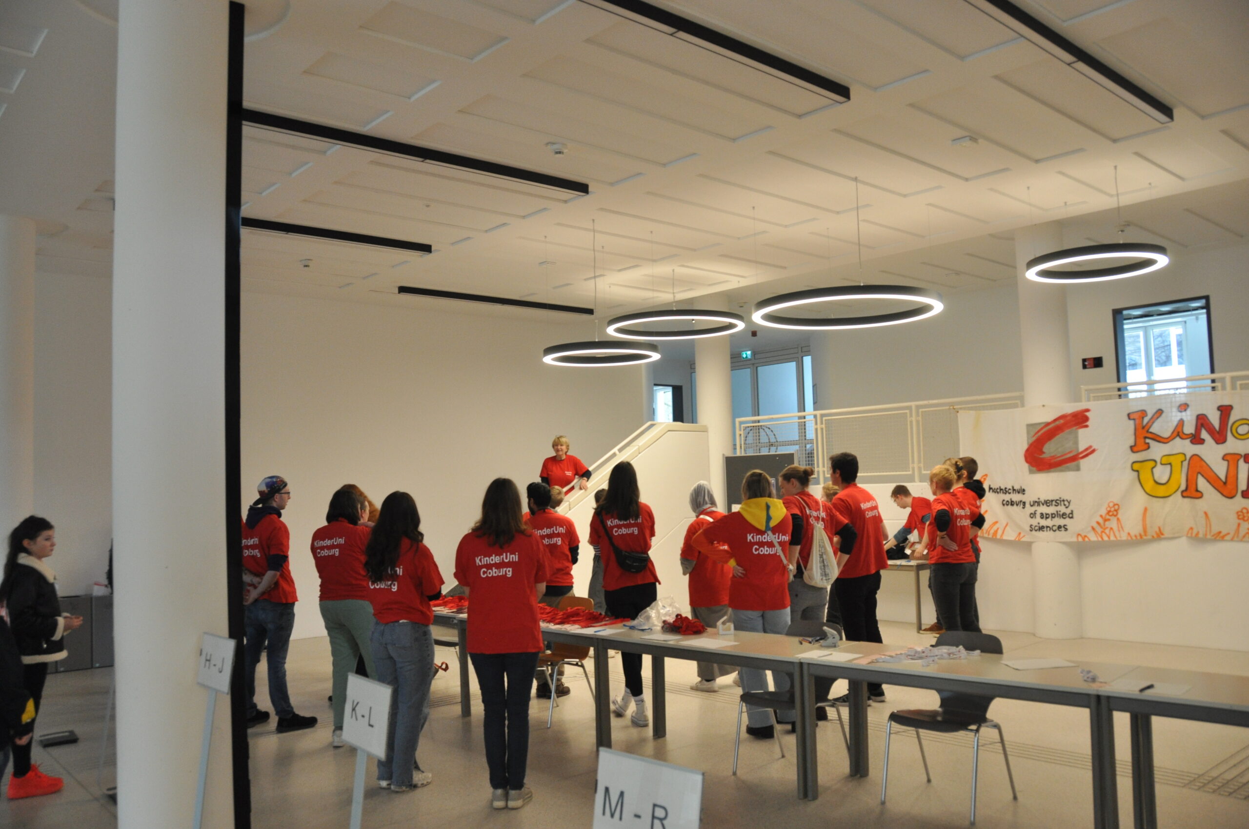 A group of people in red shirts gather in a spacious, modern room with white walls and circular ceiling lights at Hochschule Coburg. They seem to be listening to a speaker near a staircase, with signs displaying letters visible on tables.