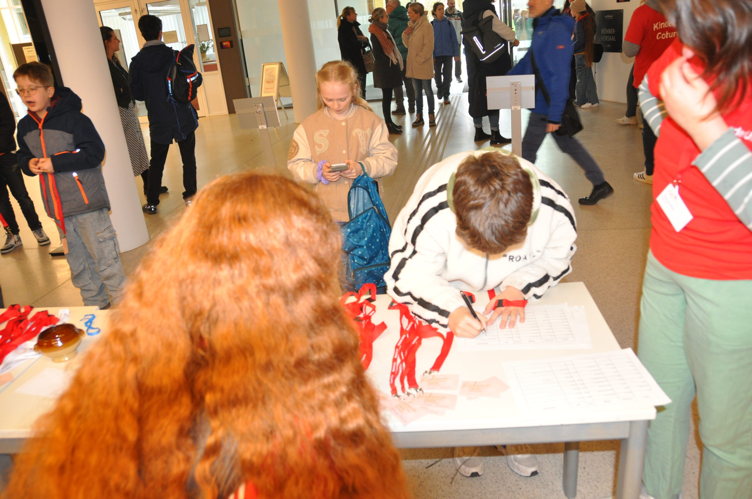 In a bustling indoor setting at Hochschule Coburg, a child scribbles on paper at a table adorned with papers and red lanyards. Nearby, a person with long red hair observes, while more people animate the background.