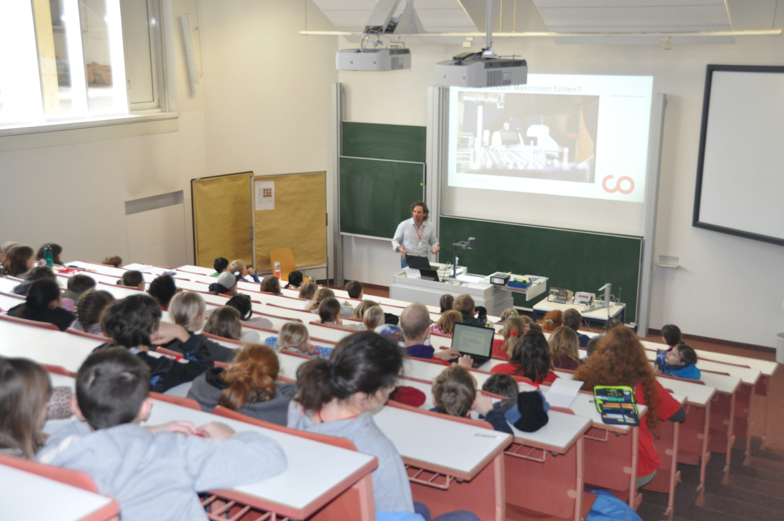 A lecturer at Hochschule Coburg stands at the front of a large classroom, presenting to a full audience of students seated in tiered rows. A presentation slide is displayed on the screen above, while some students have laptops open, eagerly taking notes.