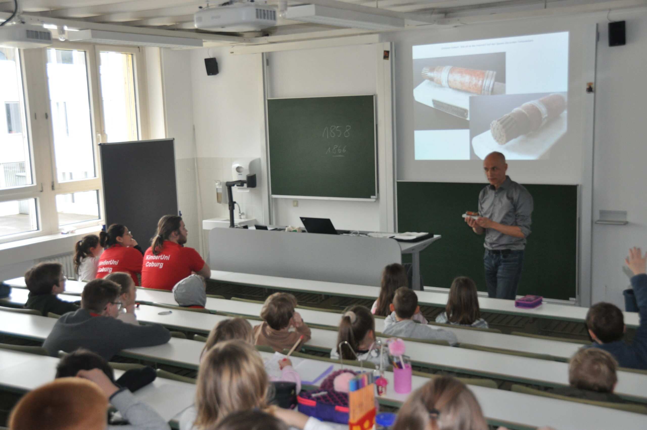 A man stands at the front of a classroom at Hochschule Coburg, giving a presentation to a group of children seated in lecture-style rows. The children are attentively listening, some wearing red shirts, as a projector displays an image of a tool on the screen.