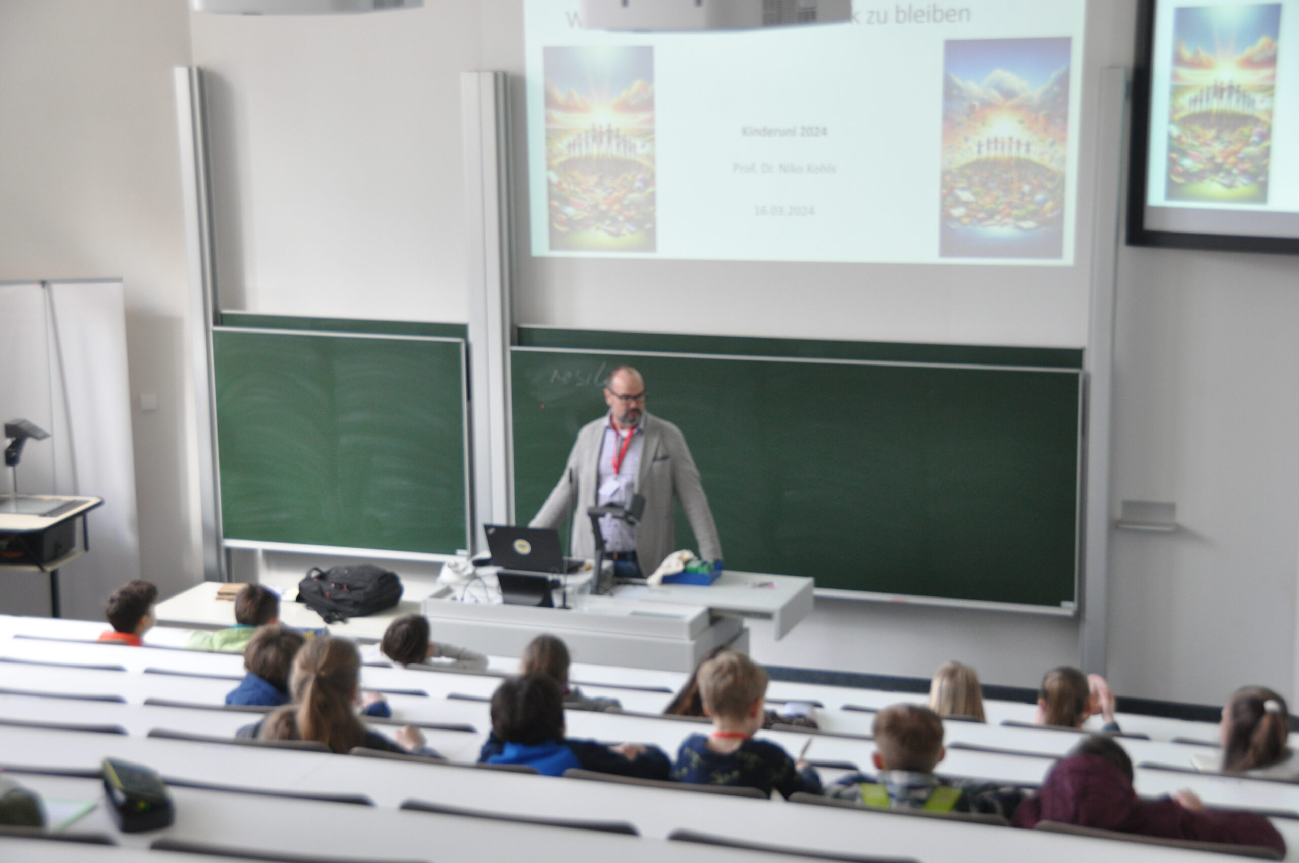 A lecturer at Hochschule Coburg stands at a podium in a large lecture hall, addressing students seated in tiered rows. A presentation glows on screens behind him, while the room, with its green chalkboards and bright lighting, fosters an atmosphere of academic engagement.