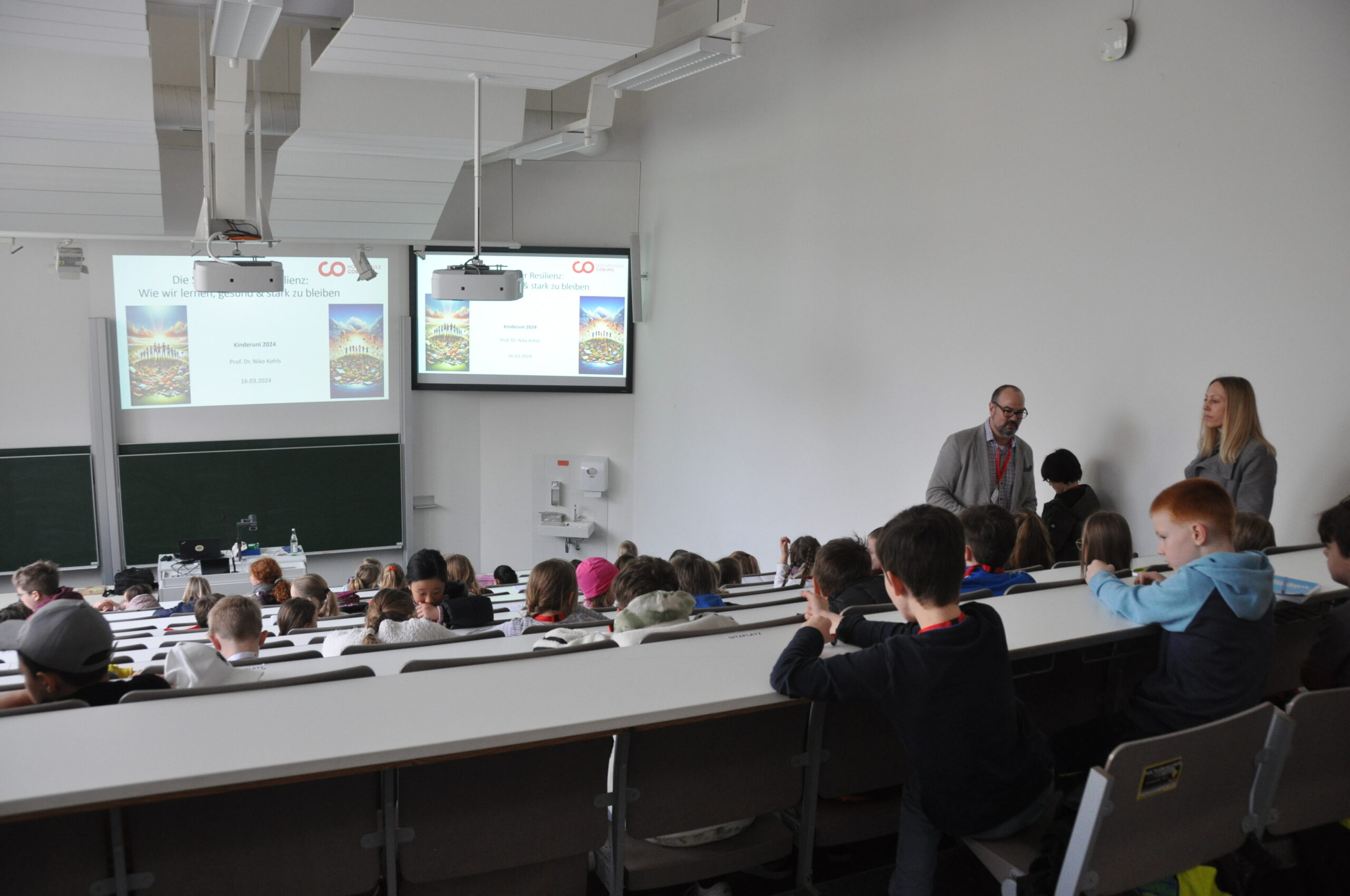 A lecture hall at Hochschule Coburg is filled with students sitting in tiered seating. At the front, two adults stand near a large screen displaying a presentation titled "We All Have a Responsibility to Listen." Participants are engaged and attentive.