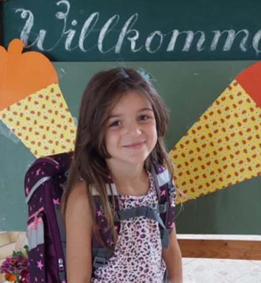 Ein junges Mädchen mit langen Haaren, einem gemusterten Kleid und einem großen Rucksack lächelt vor einer grünen Tafel mit der Aufschrift „Willkommen“. Die Tafel, geschmückt mit zwei großen Papiertüten, weist auf ihren Weg zur Hochschule Coburg hin.