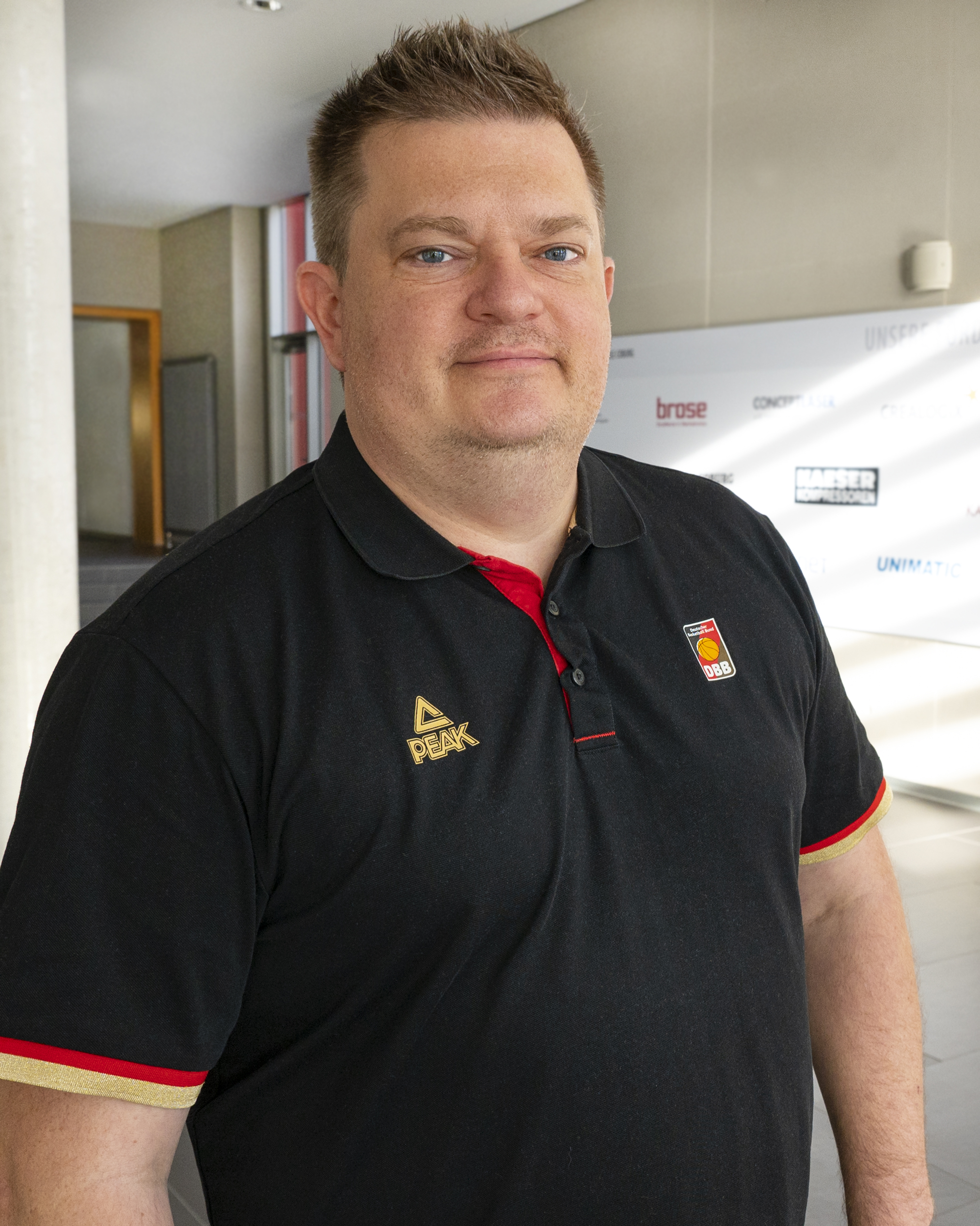 Ein Mann mit kurzen Haaren, einem schwarzen Poloshirt mit roten und goldenen Akzenten und einem Logo steht im modernen Flur der Hochschule Coburg. Im Hintergrund ist eine mit Logos geschmückte Wand und eine teilweise sichtbare offene Tür zu sehen.