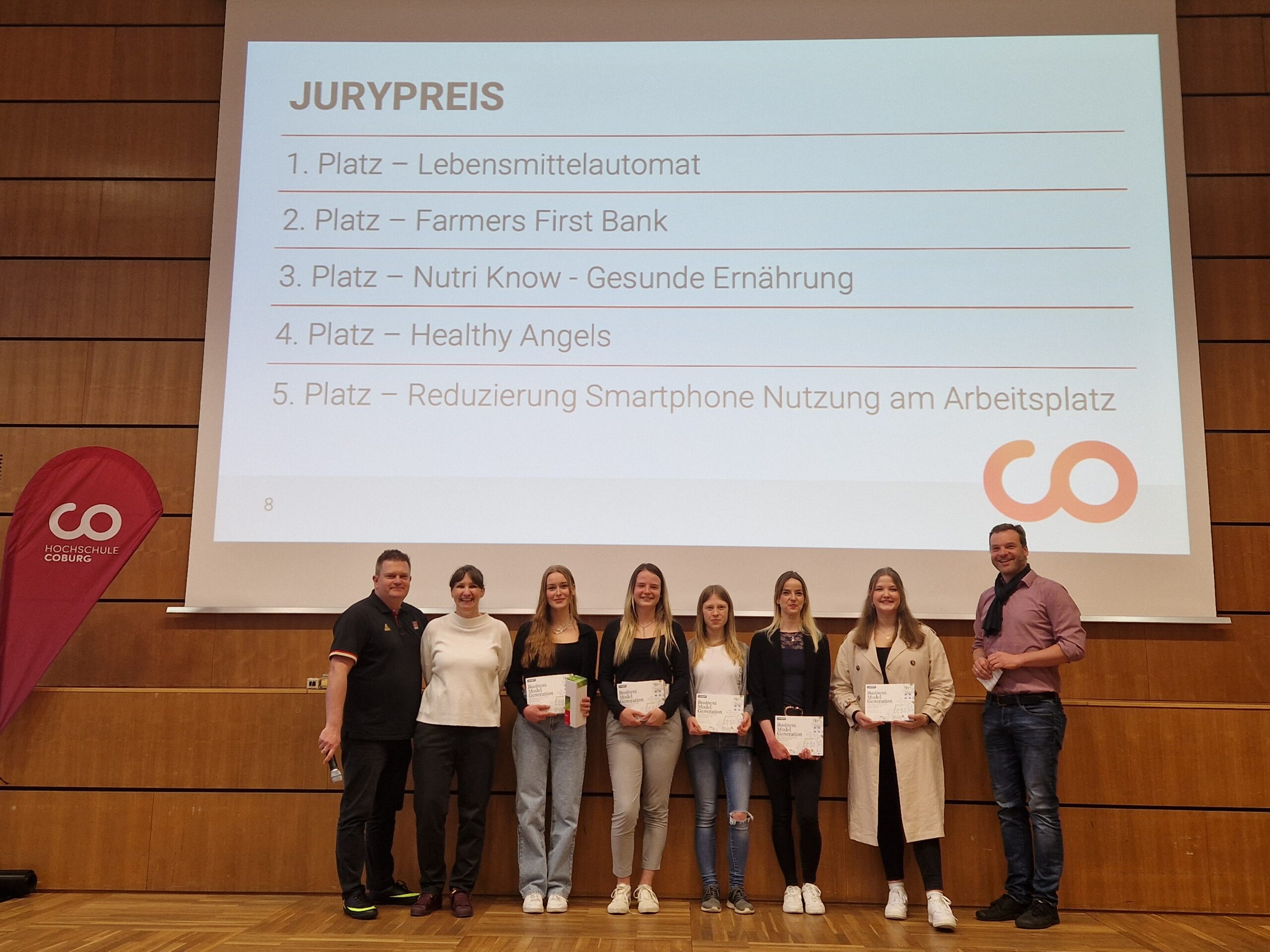 A group of seven people from Hochschule Coburg stands proudly on a stage holding certificates. Behind them, a screen displays a list of award winners under the title "JURYPREIS." On the left, a red banner adorned with a white logo adds to the celebratory atmosphere.