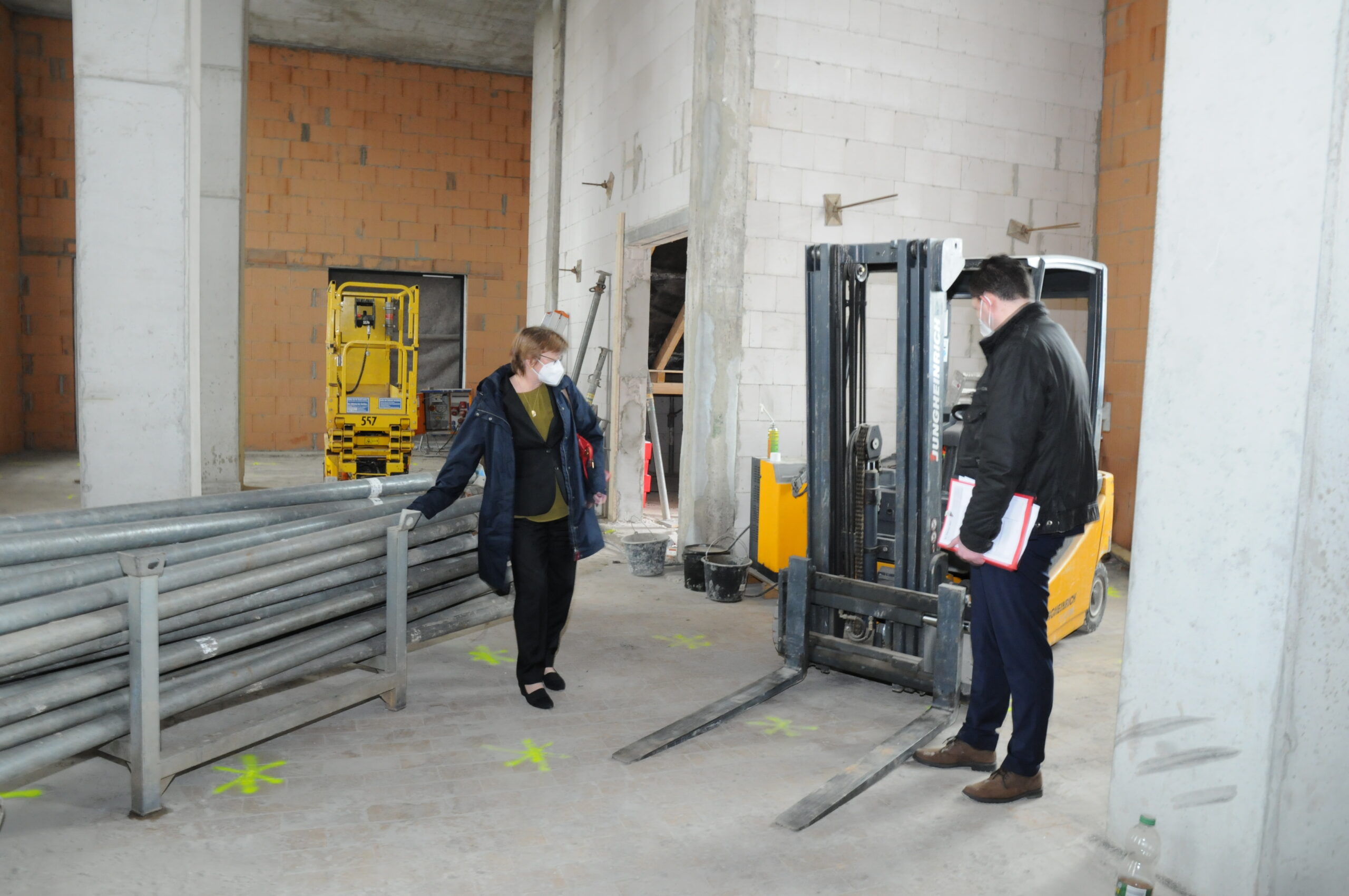 Zwei Personen inspizieren auf einer Baustelle einen Gabelstapler. Eine Person steht neben einem Stapel Metallträger, die andere hält ein Notizbuch der Hochschule Coburg. Der Bereich ist mit gelben Kreuzen markiert und hat unfertige Wände. Beide tragen Jacken, eine Person trägt eine Maske.