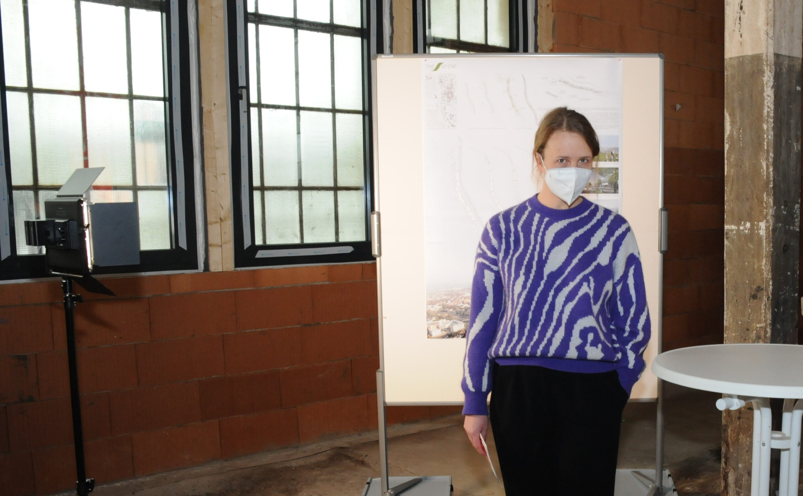 Eine Person in einem gemusterten blau-weißen Pullover und einer Gesichtsmaske steht vor einer Präsentationstafel an der Hochschule Coburg, einem industriell anmutenden Raum mit großen Fenstern als Hintergrund.